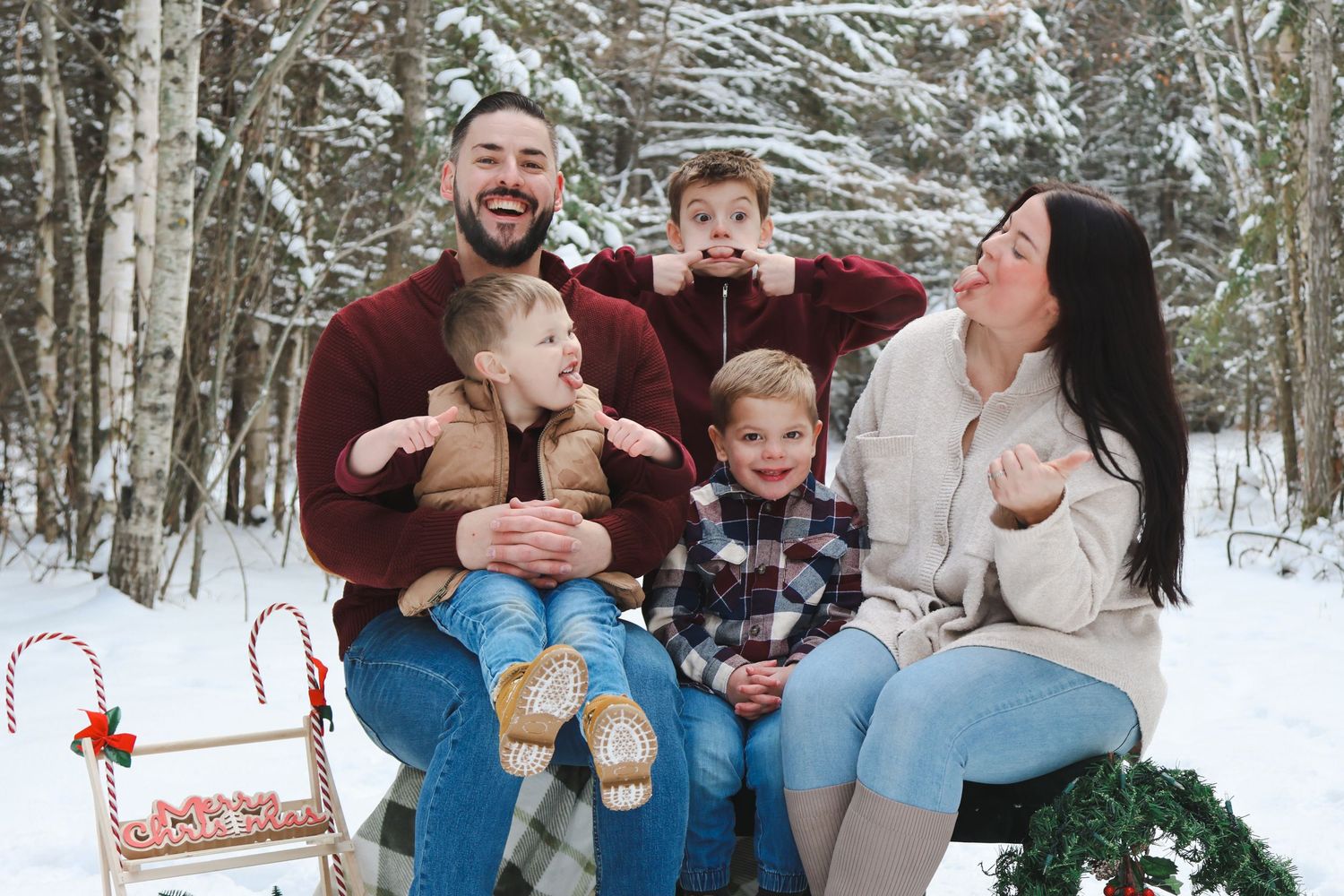 Playful family of five makes funny faces in snowy winter forest with Christmas sled decor.