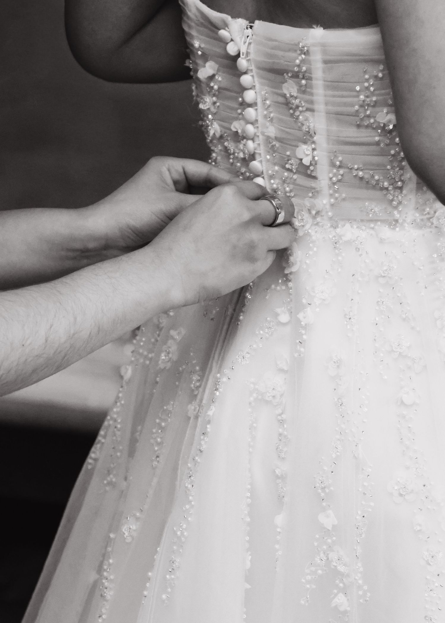 Black and white close-up of hands fastening buttons on the back of a wedding dress.