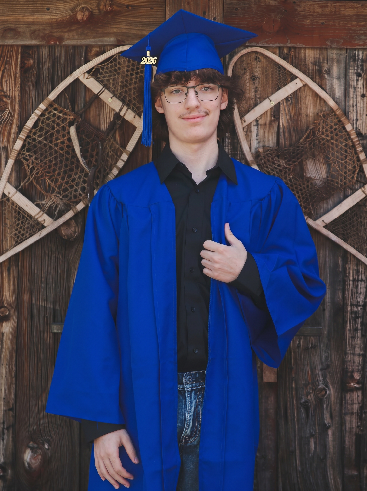 Young graduate in blue cap and gown giving thumbs up, posing in front of rustic wooden wall with snowshoes.