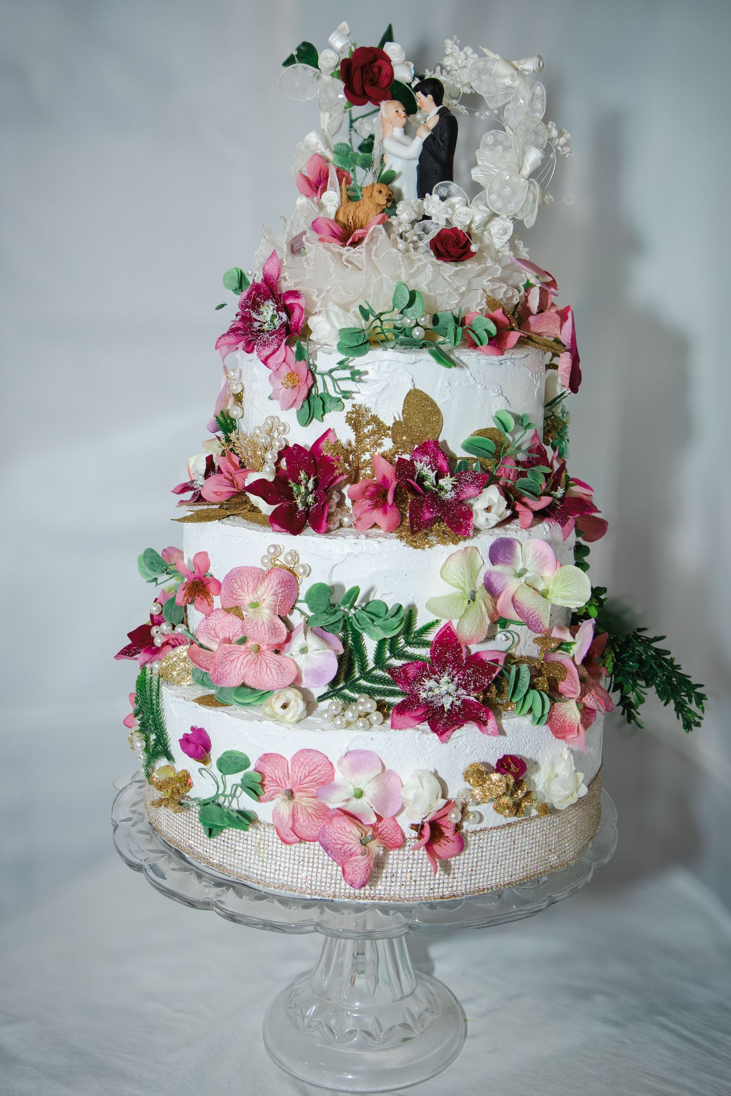 Three-tier white wedding cake adorned with colorful floral decorations and bride and groom topper on crystal cake stand.