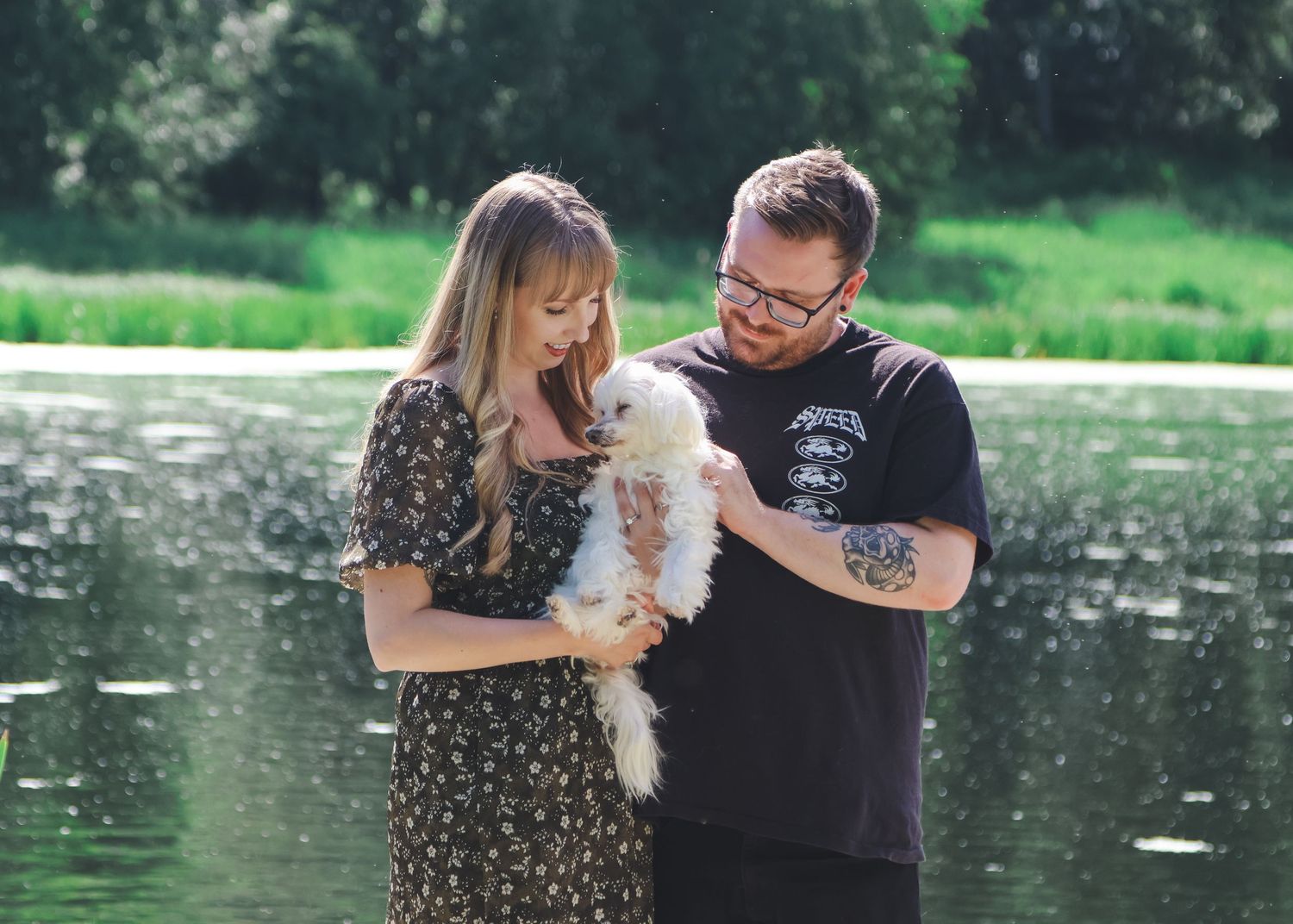 Couple standing by a lake holding a small white fluffy dog, surrounded by lush green trees on a sunny day.