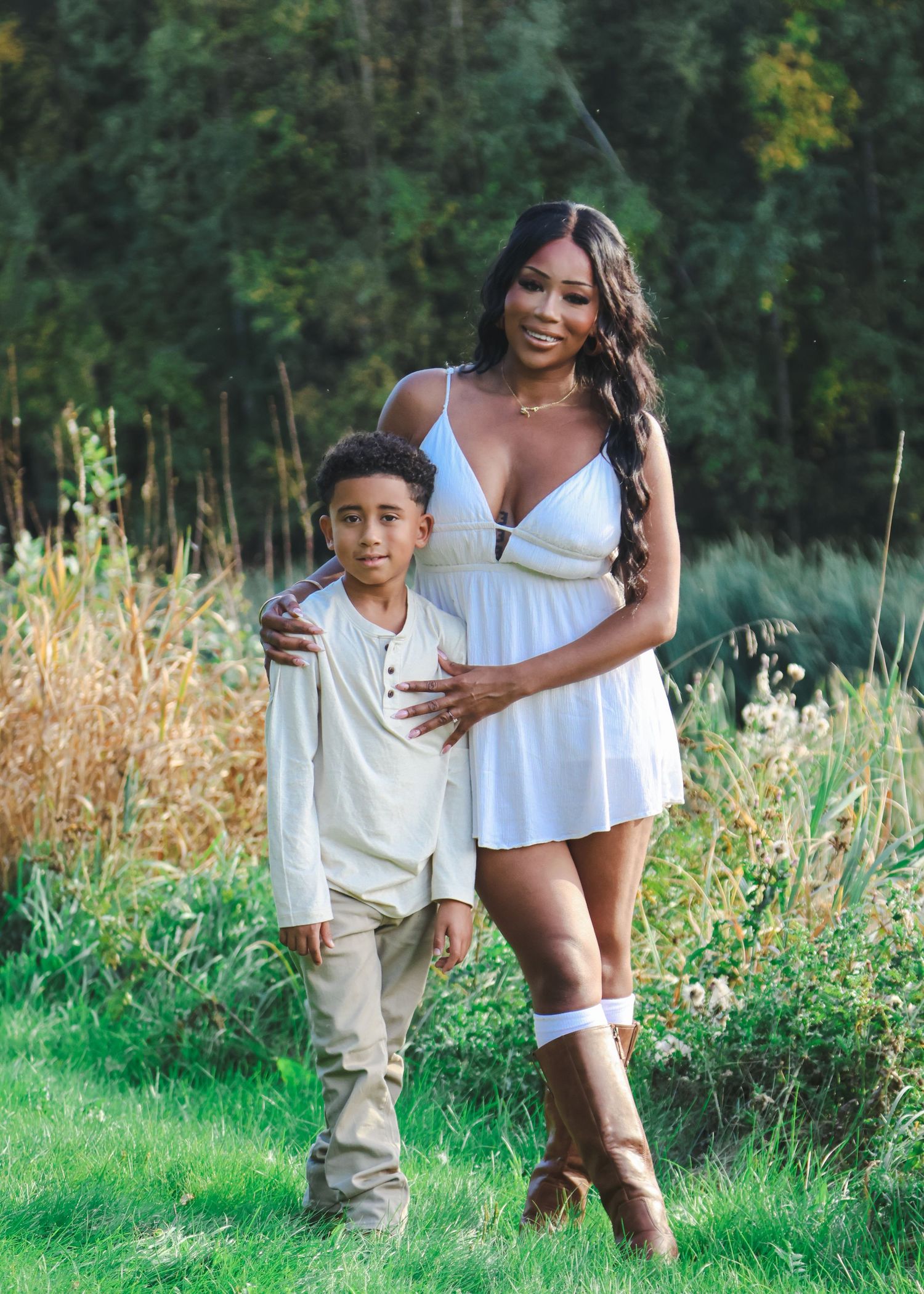 A woman in a white dress and a young boy in cream outfit pose together in a lush green outdoor setting.