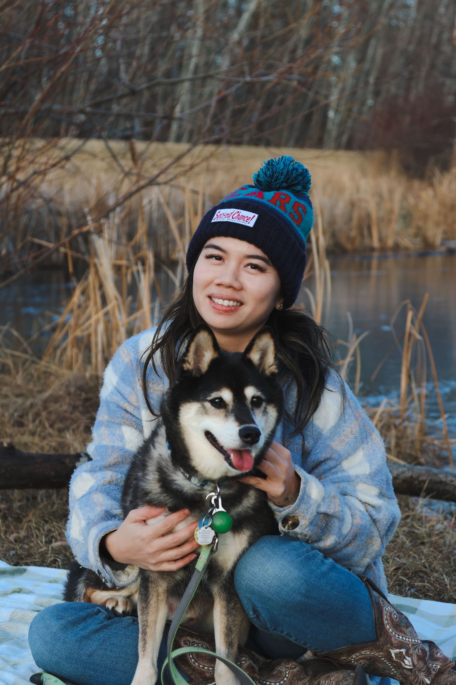 Smiling young woman in a pom-pom beanie hugging her Husky dog outdoors near a winter pond.