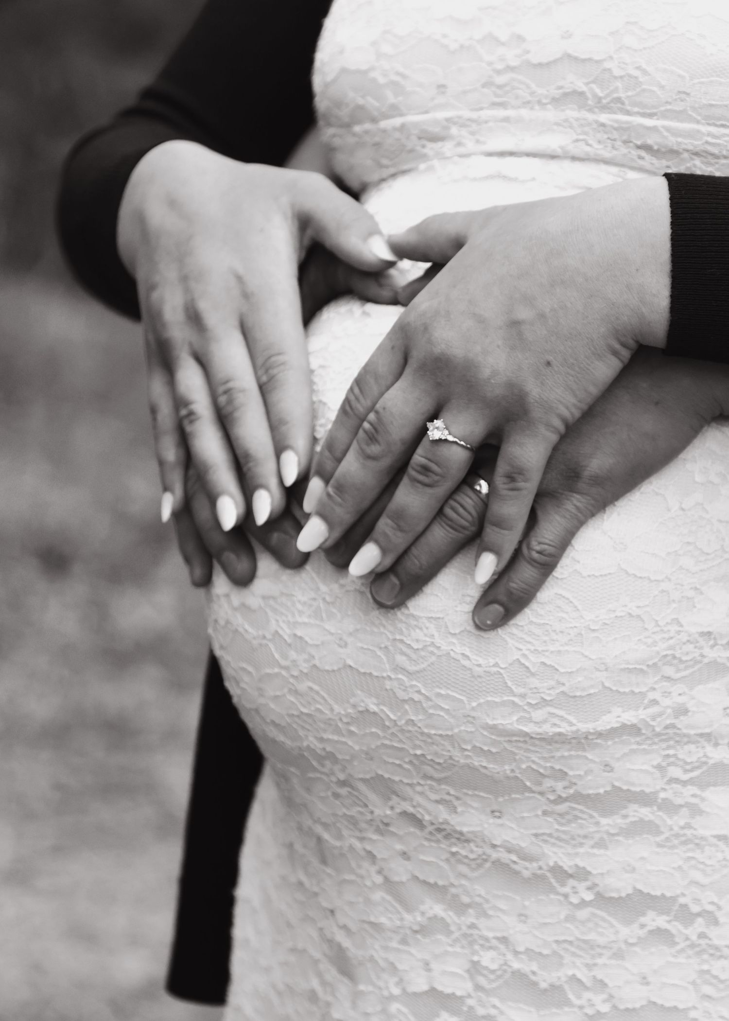 Black and white close-up of hands on pregnant belly in white lace dress with engagement ring.