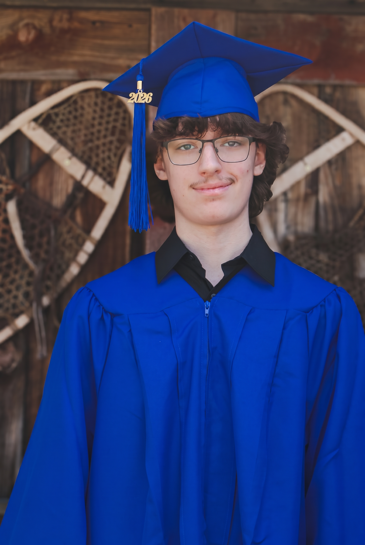 Young man in blue graduation cap and gown smiling for senior portrait against rustic wooden background.
