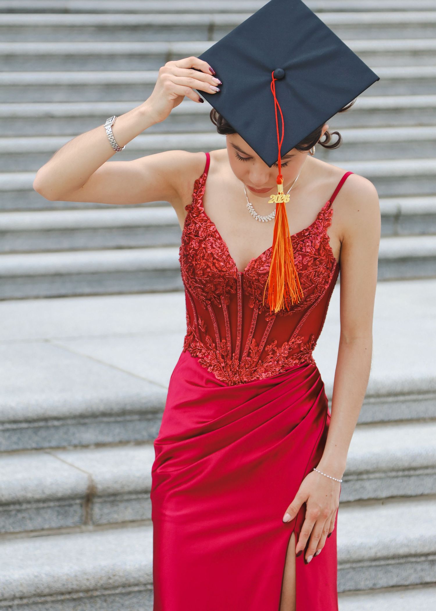 Elegant female graduate in red sequin gown adjusting black mortarboard cap on marble steps.