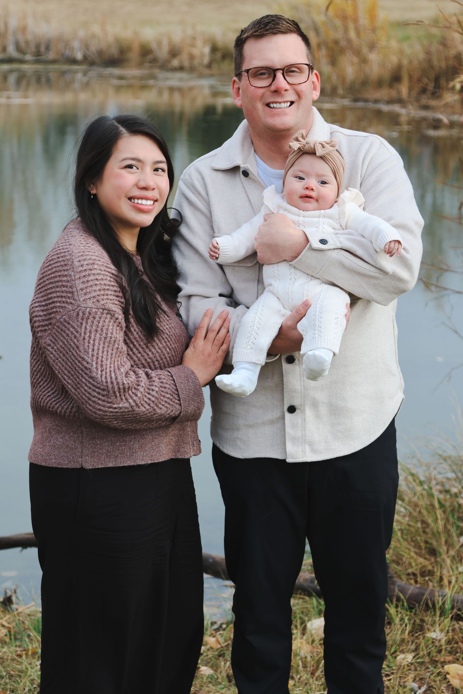 Happy family of three stands by pond, man holding baby in white while woman smiles beside them.