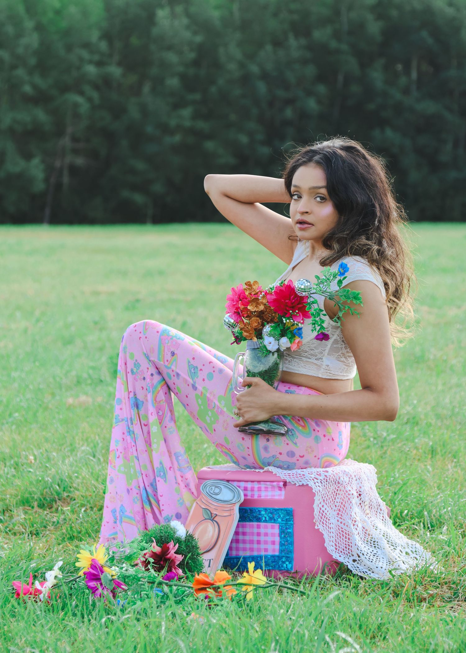 Young woman in pink floral pants holds colorful bouquet while sitting on pink box in a green field.
