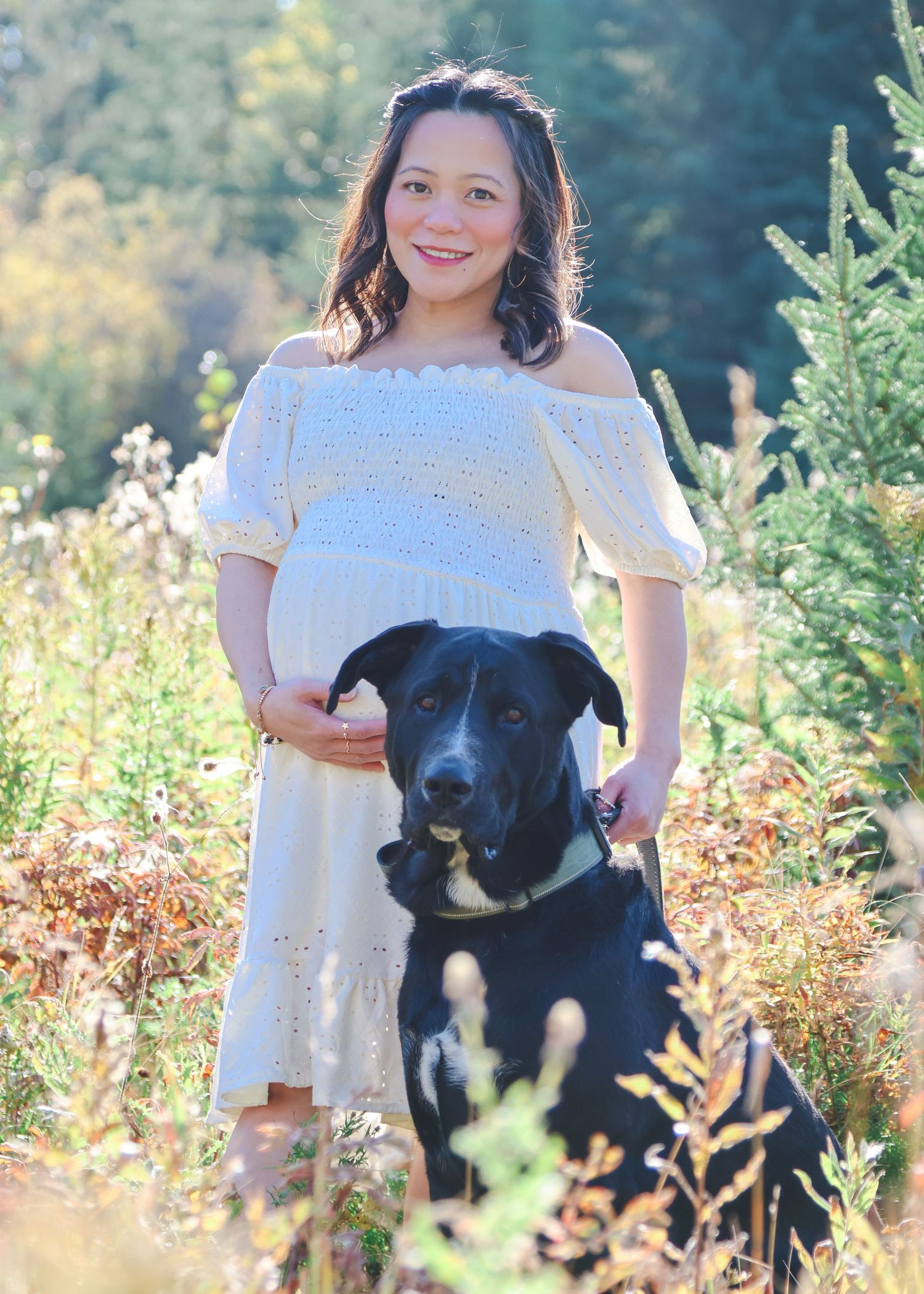 Pregnant woman in white off-shoulder dress poses with black Labrador mix in sunny outdoor field.