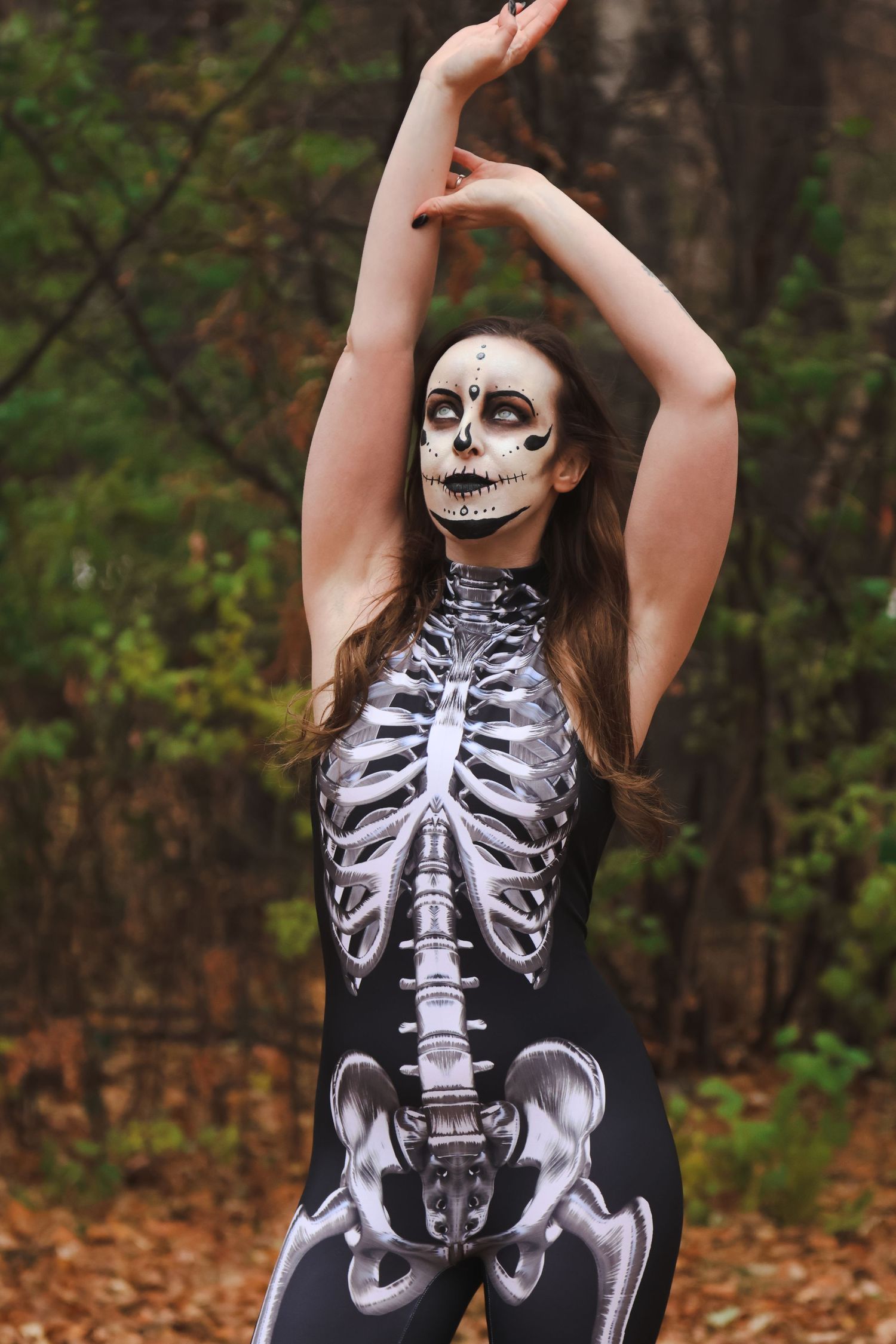 Woman in skeleton Halloween costume and skull face paint posing in autumn forest setting.