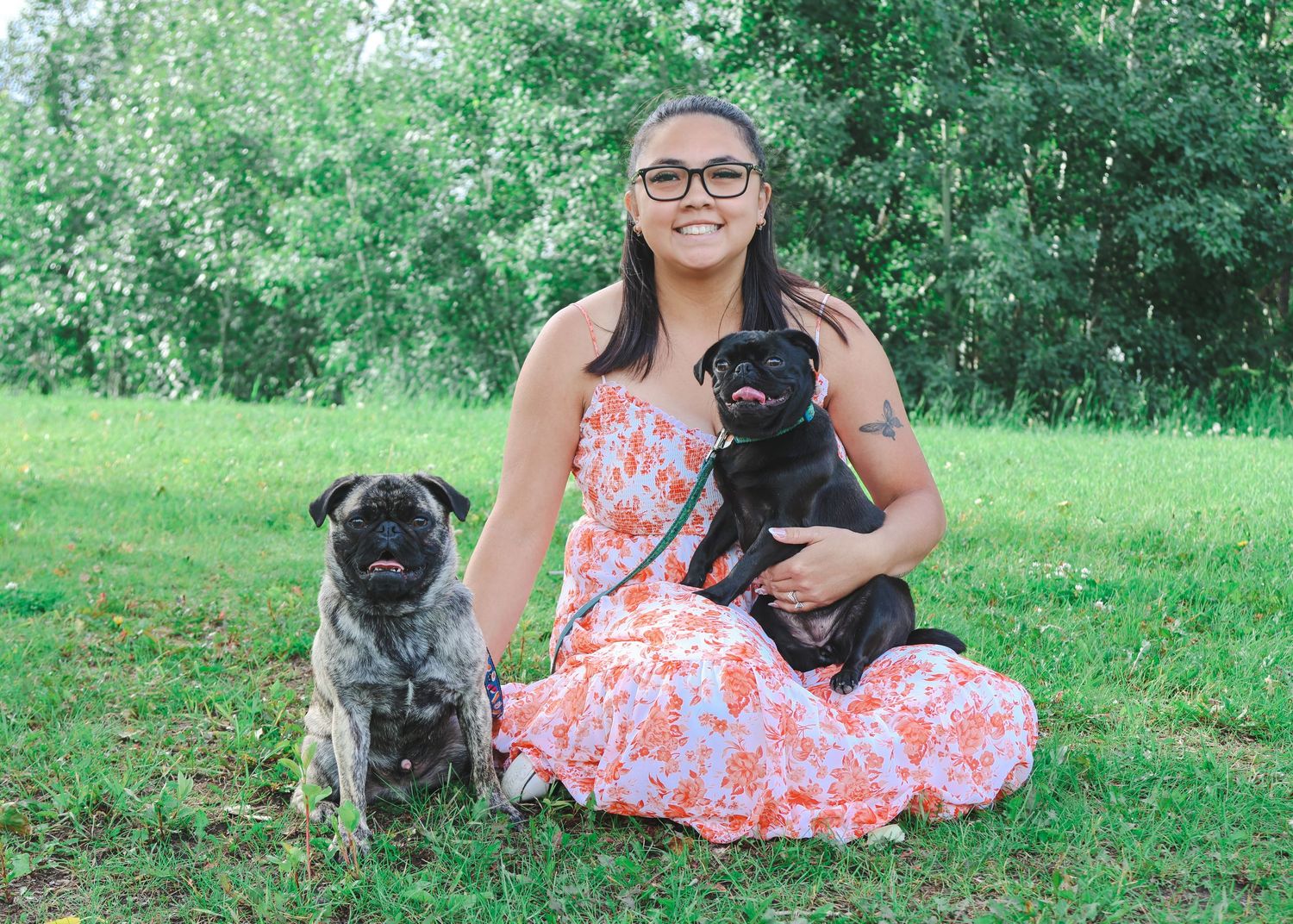 Woman in floral dress sitting cross-legged on grass with two black pugs in a park setting.