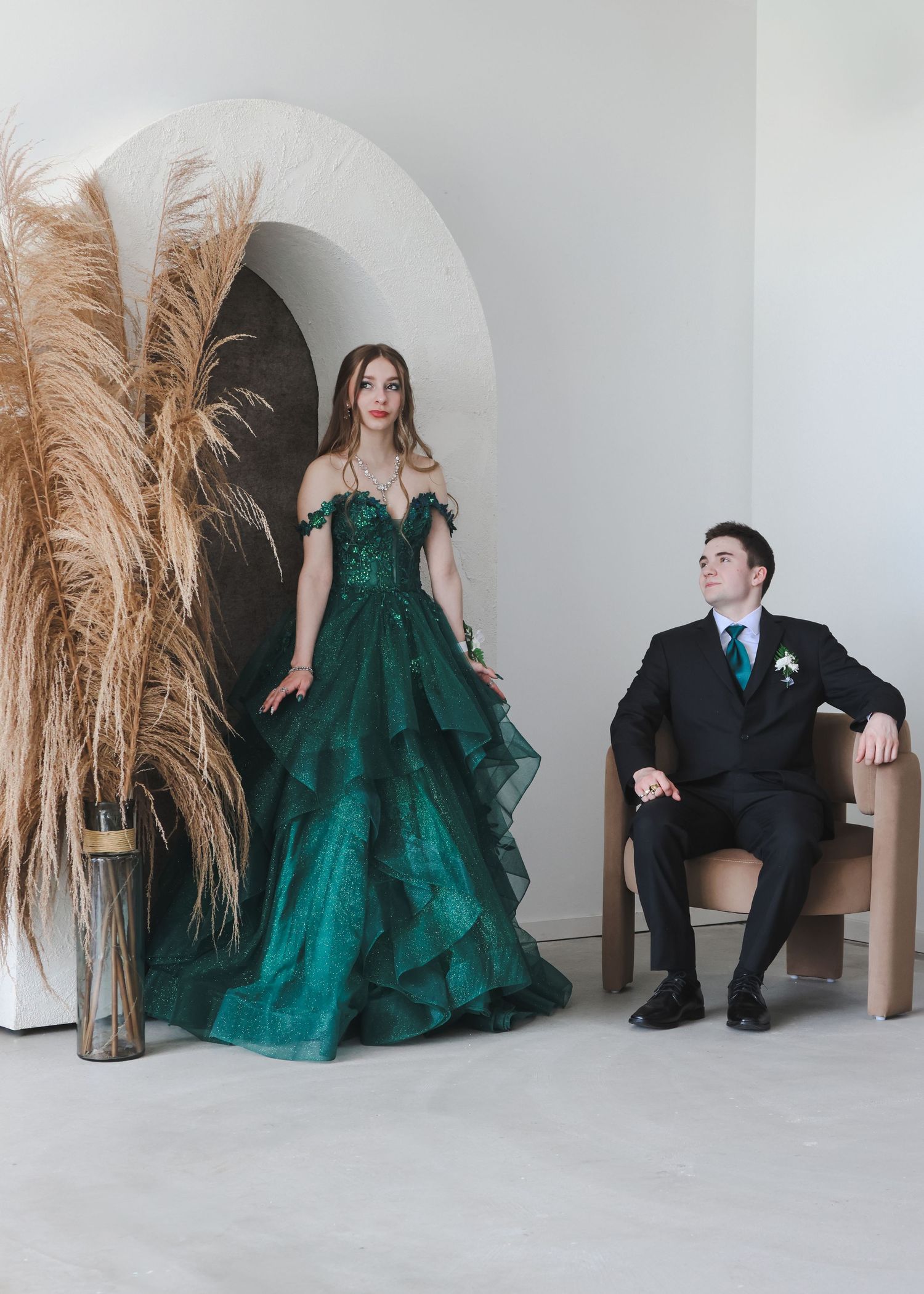 Couple in emerald green ball gown and black tuxedo posing near pampas grass and arched doorway.