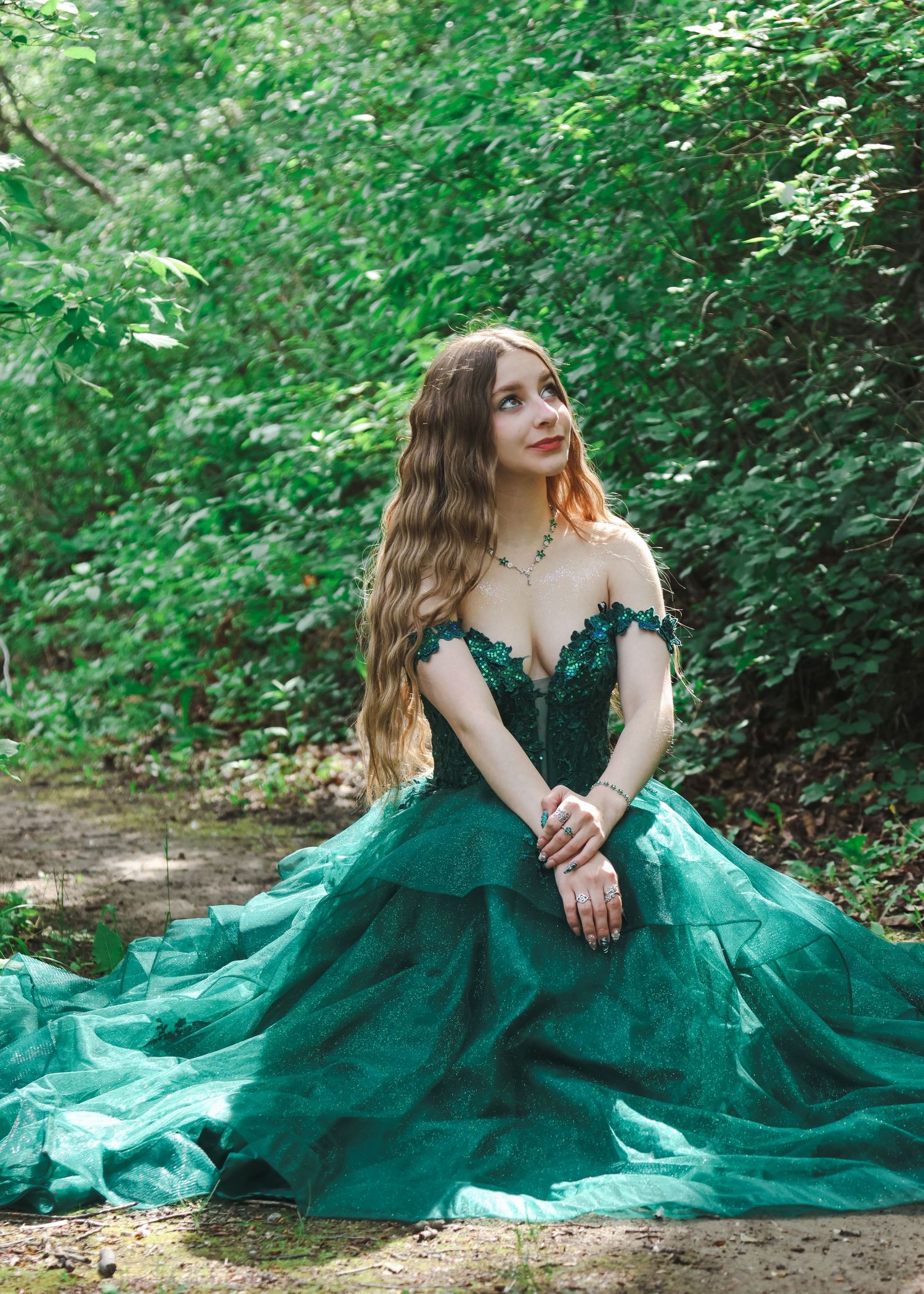 Young woman in an off-shoulder emerald green tulle gown sits gracefully in a lush green forest.