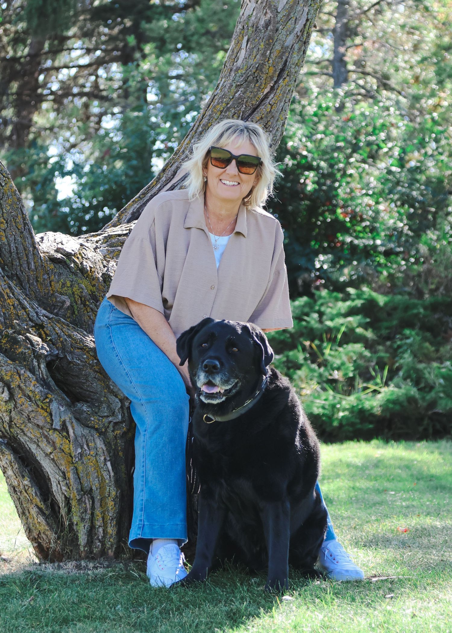 Woman with sunglasses smiling beside a large tree trunk with a black Labrador Retriever on a sunny day.