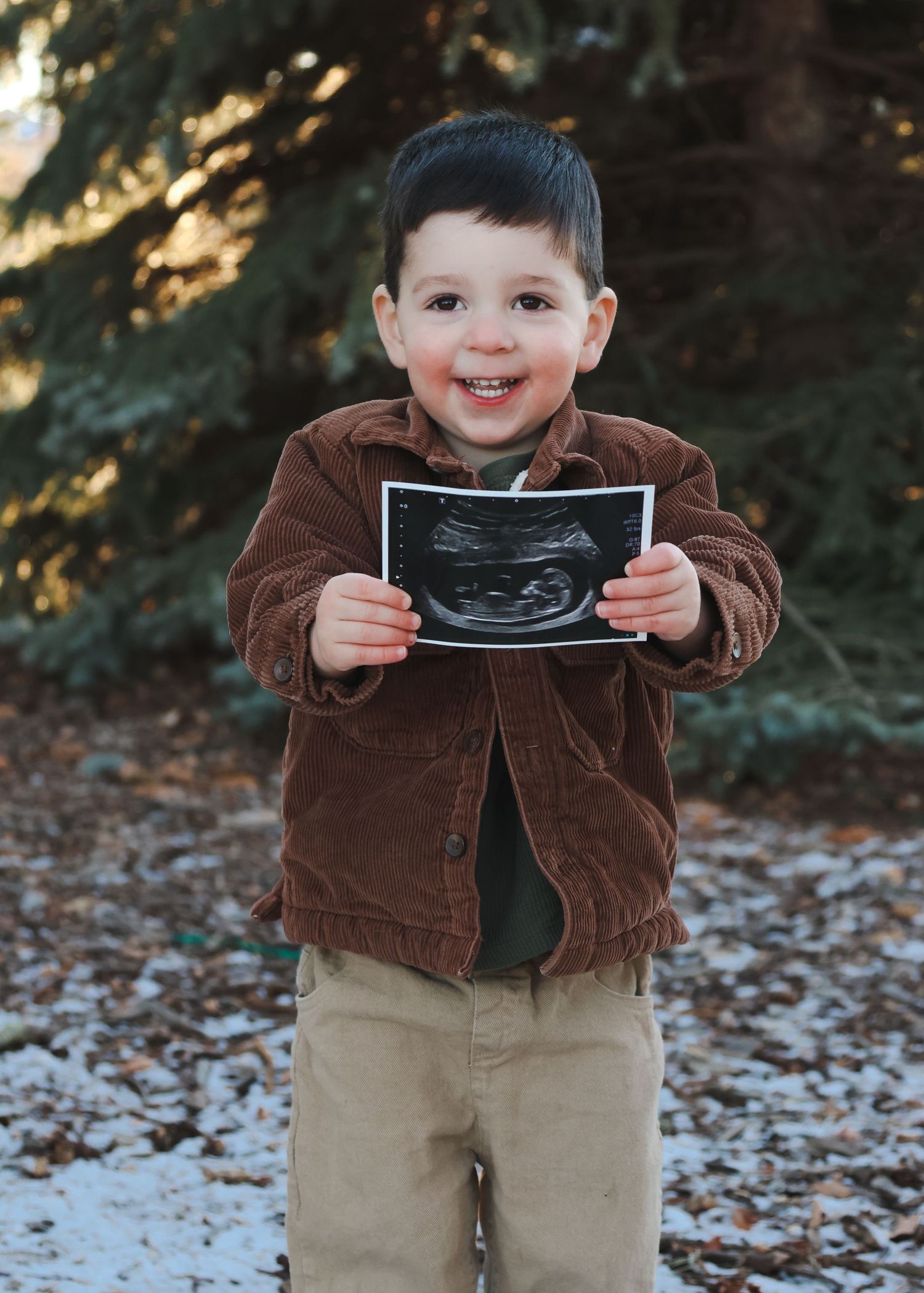 Smiling toddler boy holding ultrasound photo outdoors in winter, wearing brown corduroy jacket.