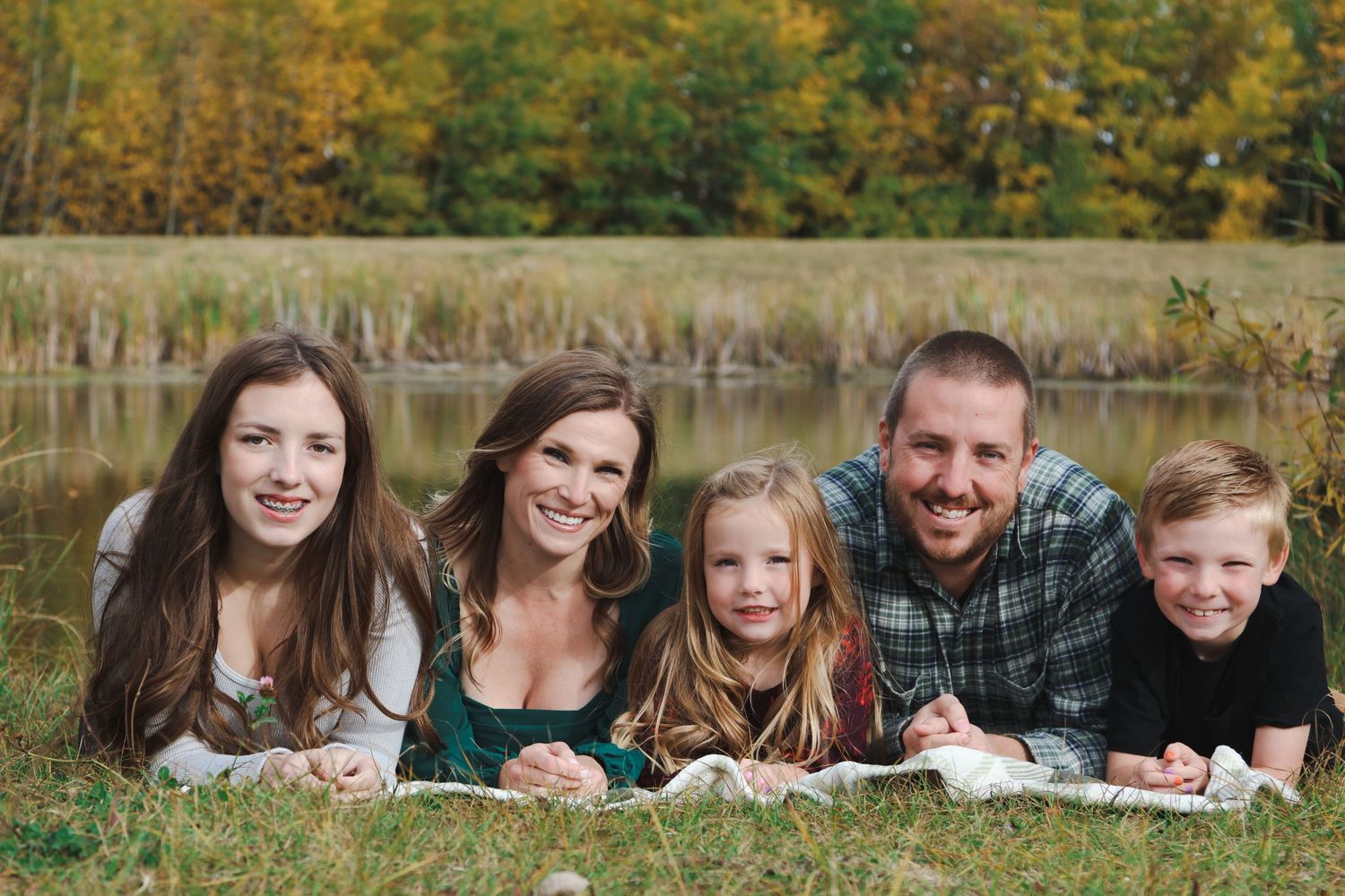 Smiling family of five poses lying on blanket in front of autumn pond with colorful foliage.