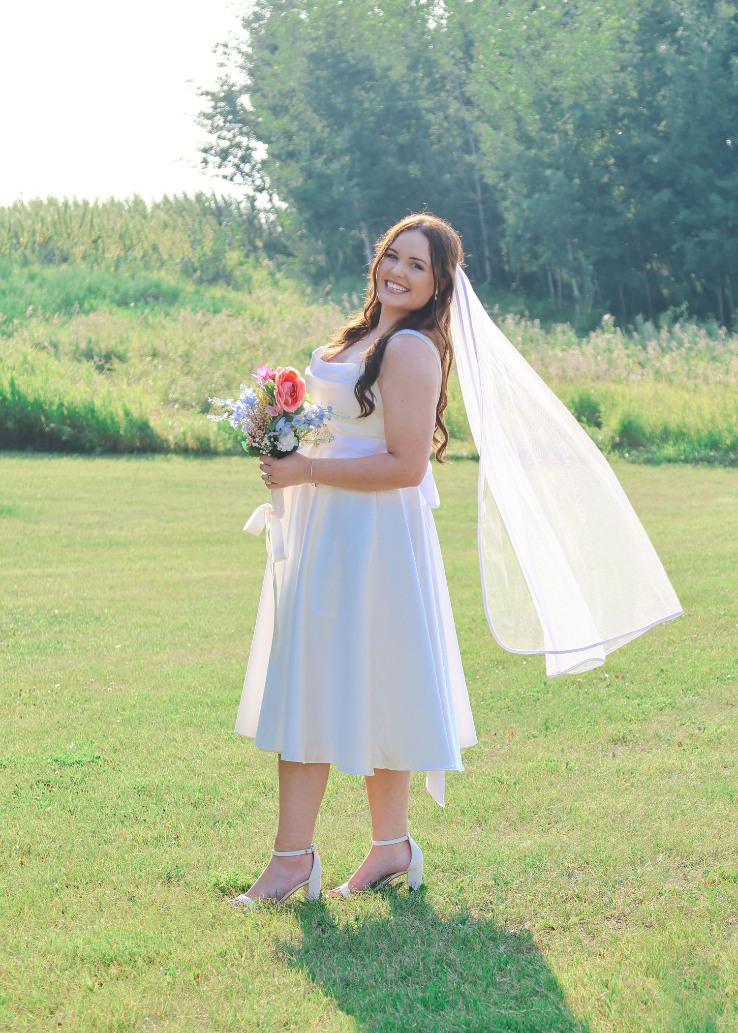 Happy bride in white midi dress holding bouquet with flowing veil in sunny green field.