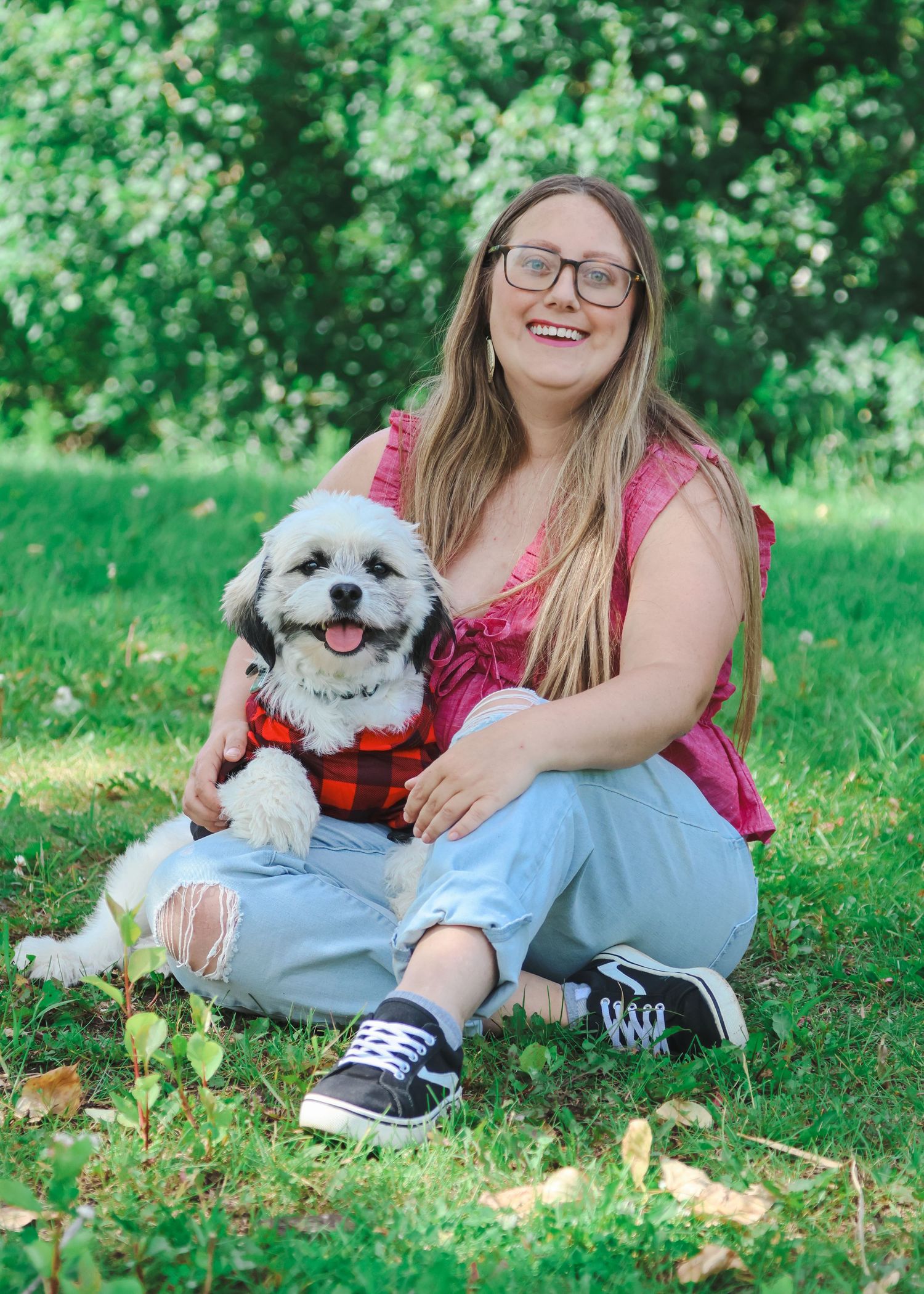 Smiling woman with glasses sitting on grass holding a small white fluffy dog in a red plaid outfit.