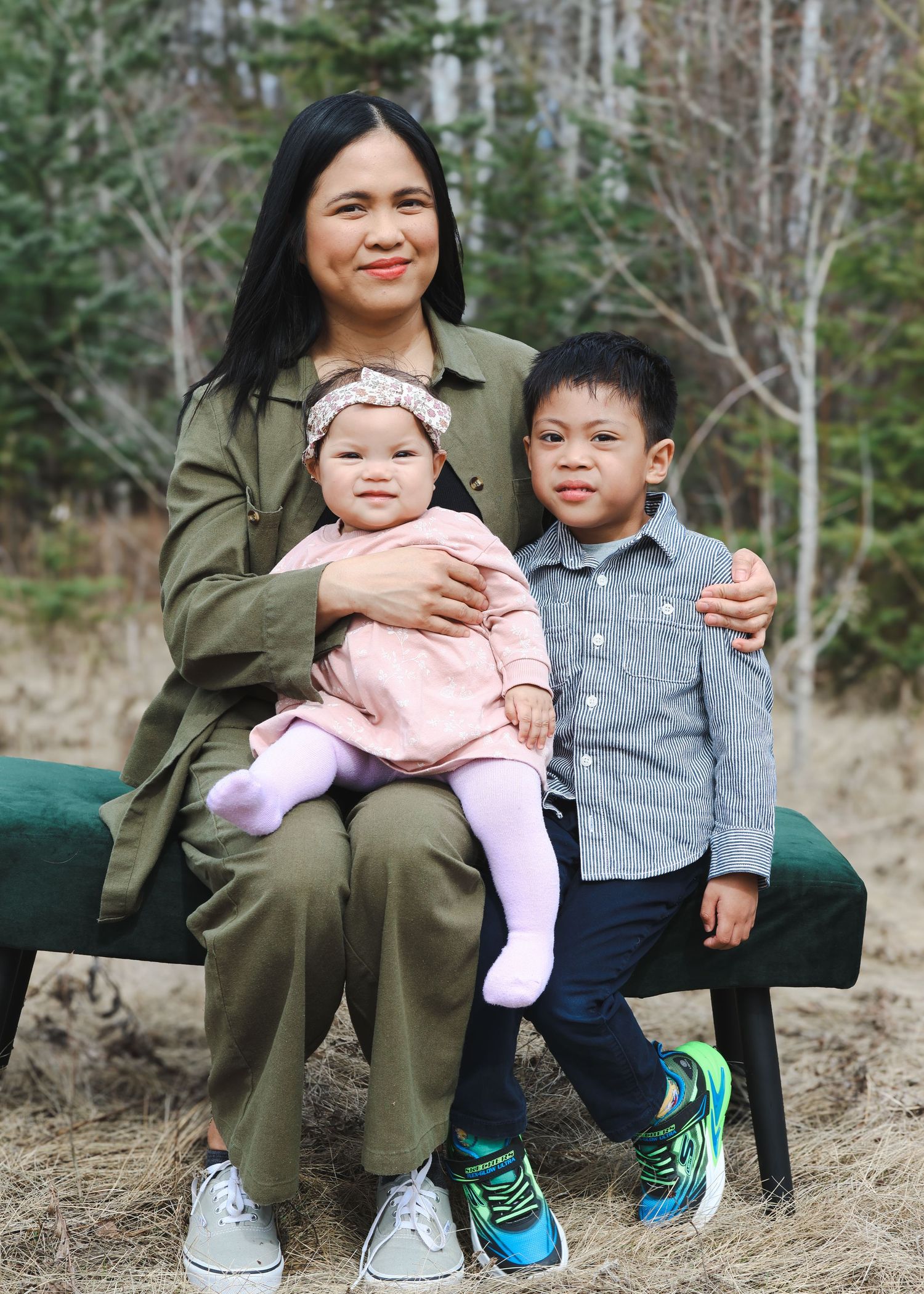 Mother in olive green sits on bench with baby daughter and young son in forest setting.