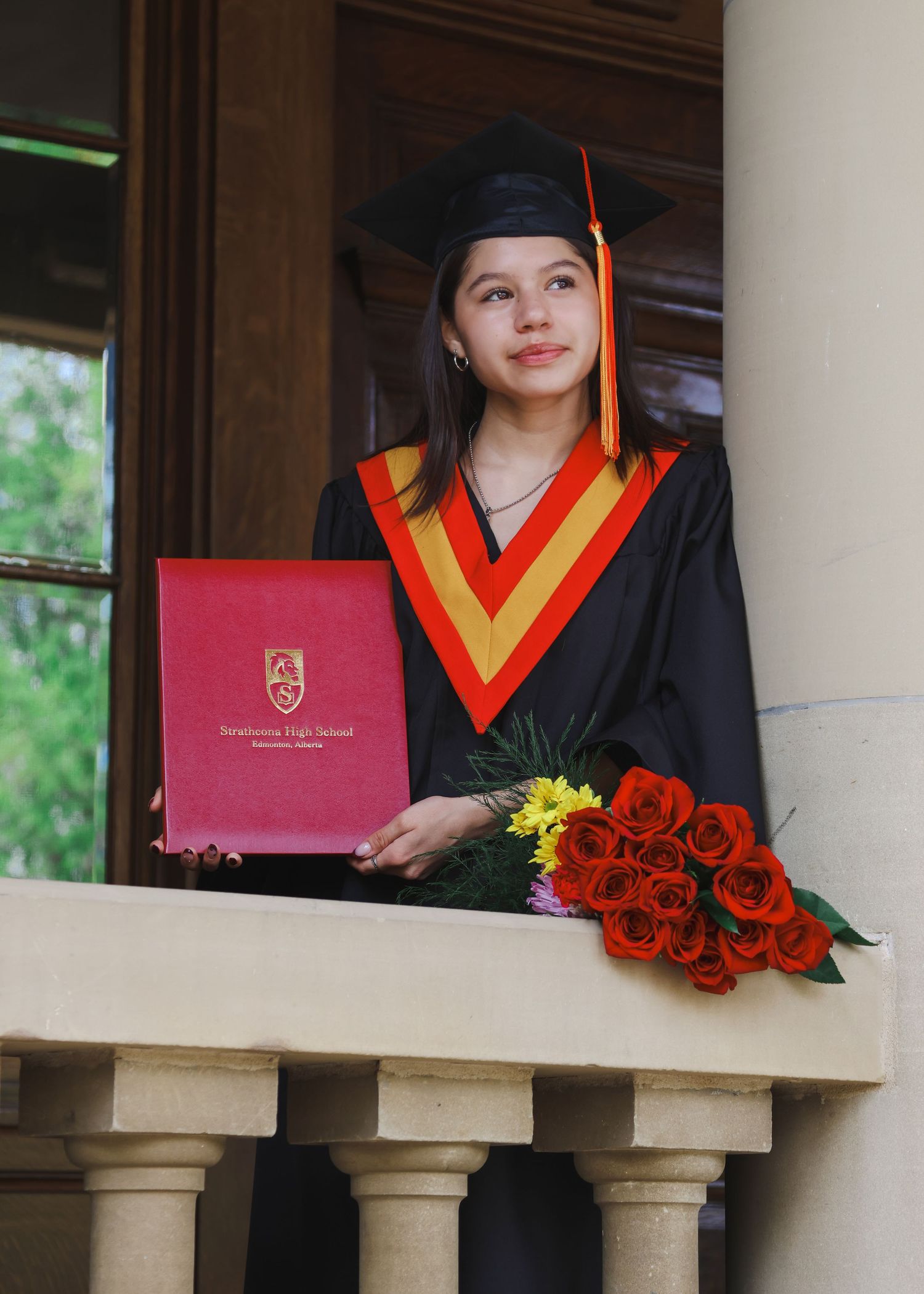 Graduate in black cap and gown holding red diploma and roses on stone balcony.