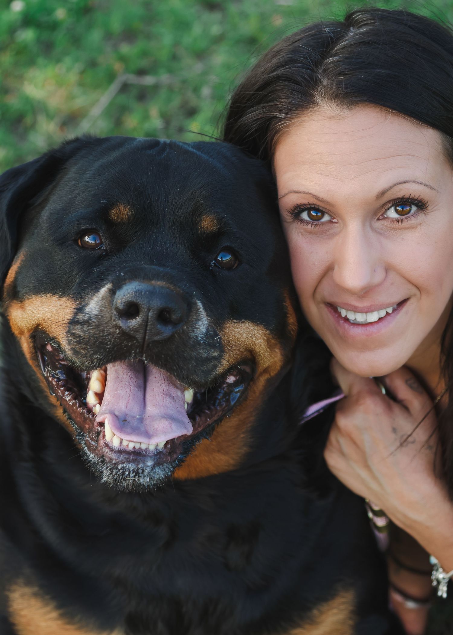 A smiling woman poses closely with a happy Rottweiler dog outdoors on green grass.