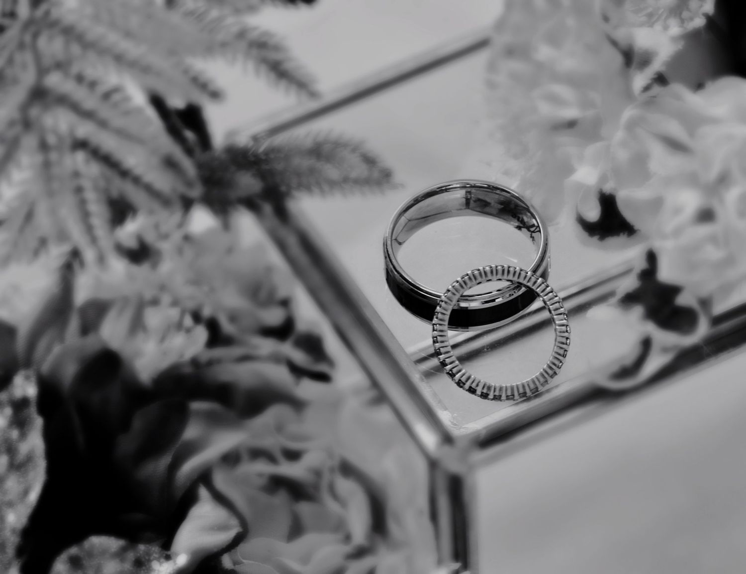 Black and white photo of wedding rings resting on a glass jewelry box surrounded by flowers.