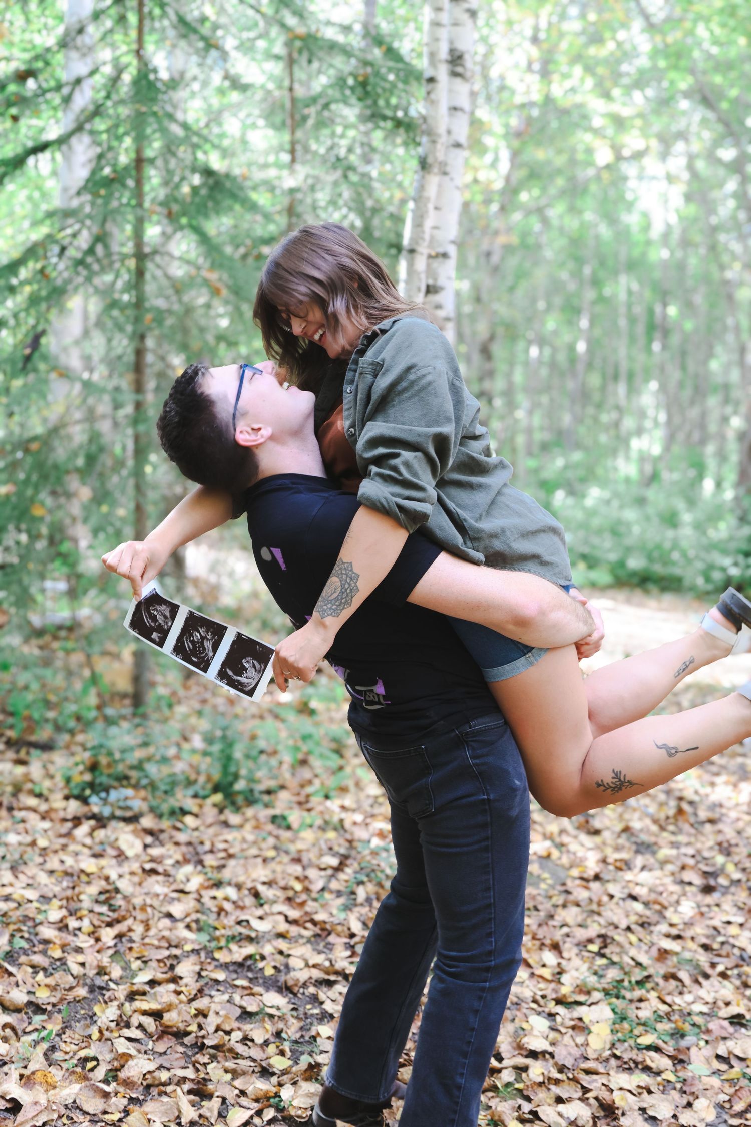 Man lifting woman in autumn forest while she holds ultrasound photos, pregnancy announcement.