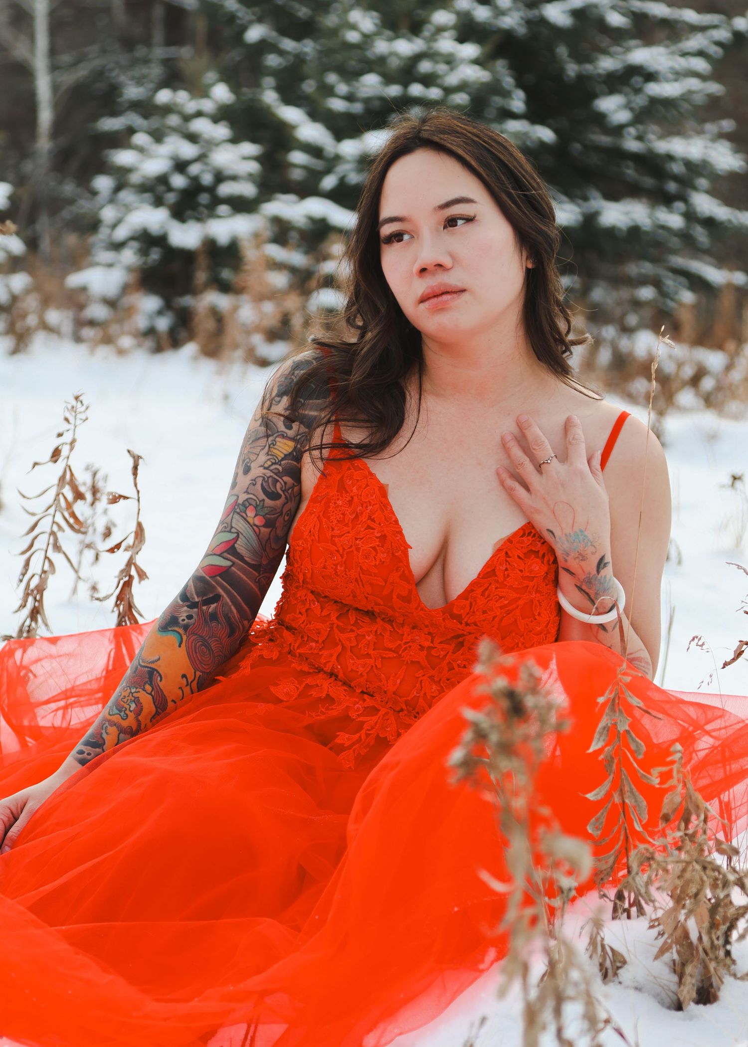 Woman in red gown with sleeve tattoos poses gracefully in snowy winter forest setting.