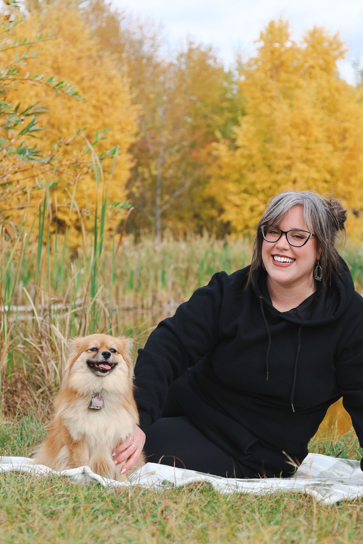 Woman in black hoodie smiling with fluffy Pomeranian dog outdoors among autumn yellow trees.
