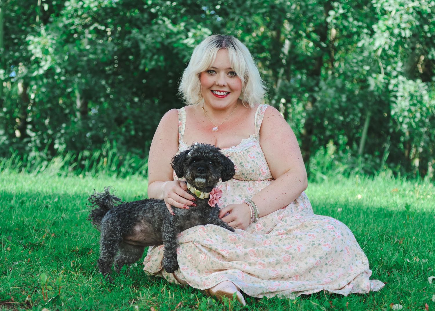 Woman in floral dress sitting on grass with two small black dogs in a lush green park setting.