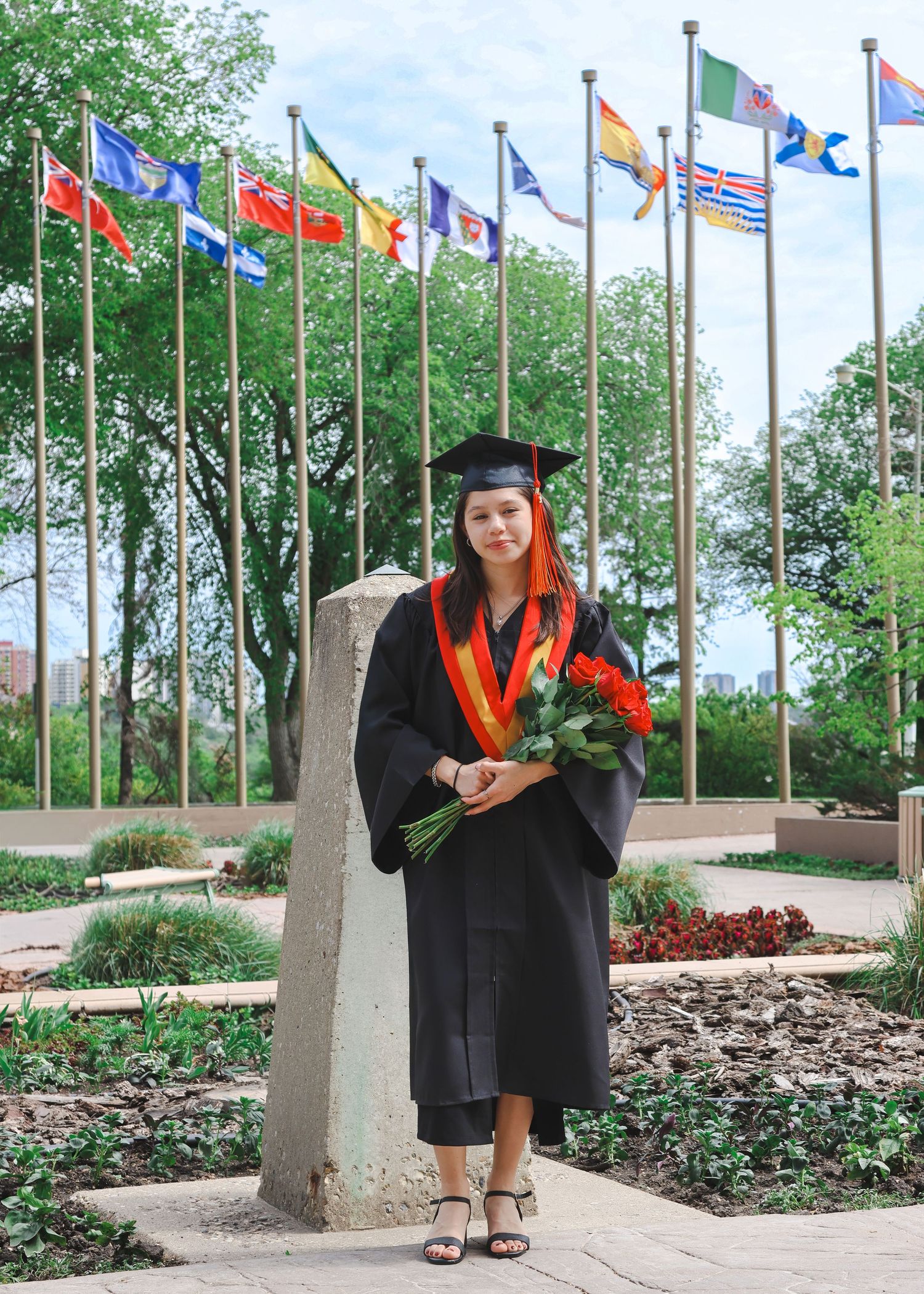 Graduate in black cap and gown with red roses standing before international flags.