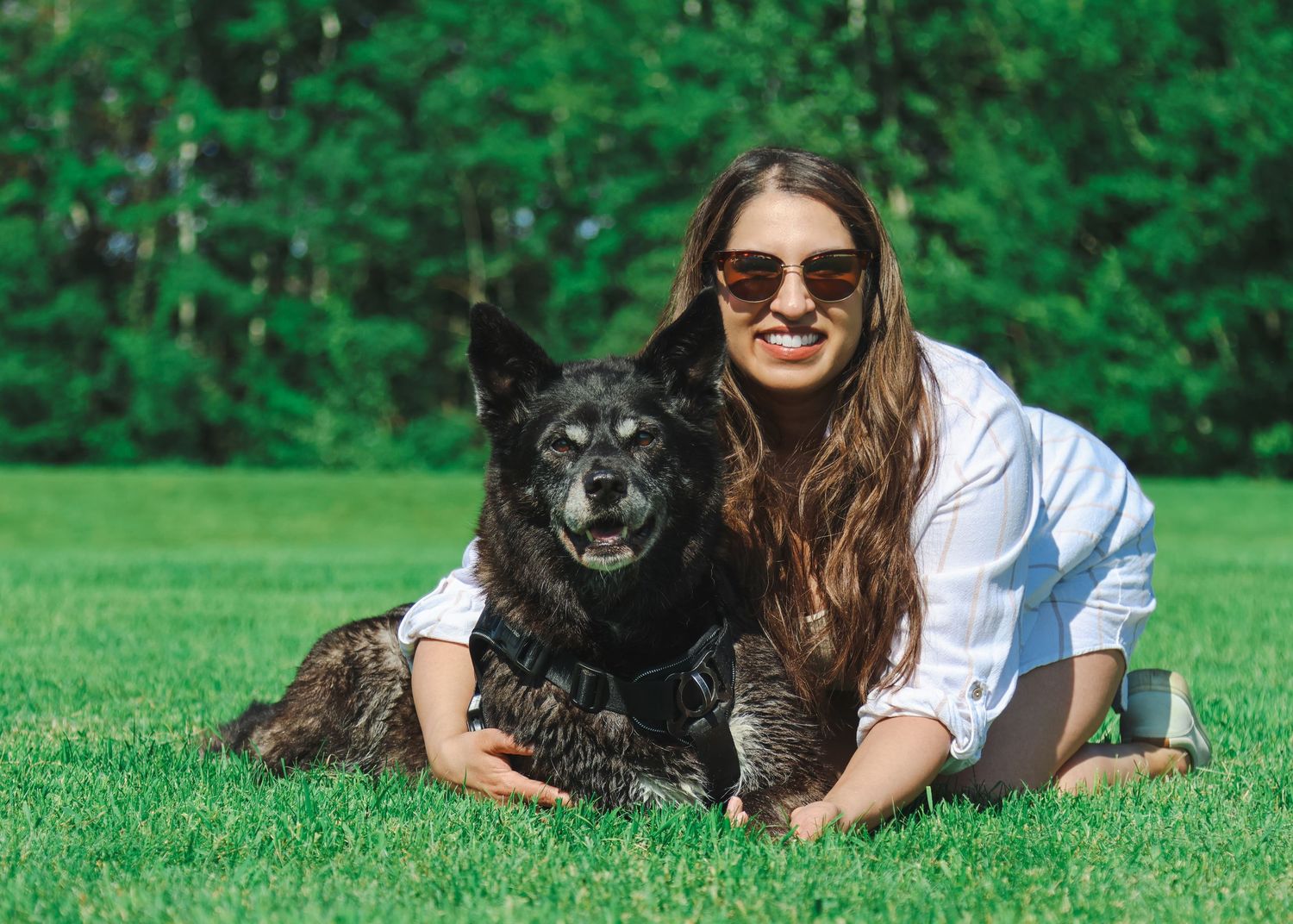 Young woman with sunglasses posing with a large black dog on green grass in a park.