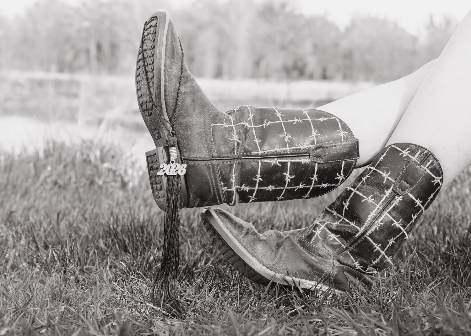 Black and white close-up of cowboy boots with 2024 graduation charm on grass.