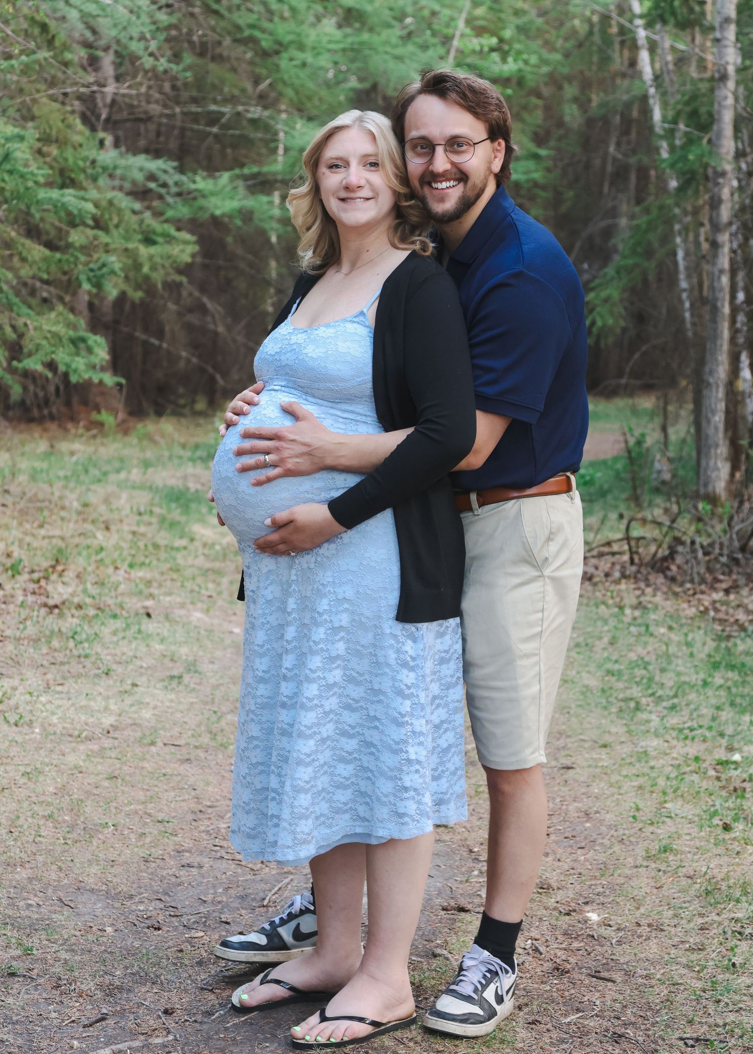 Happy expecting couple embracing outdoors, woman in blue lace dress and man in navy shirt smiling.