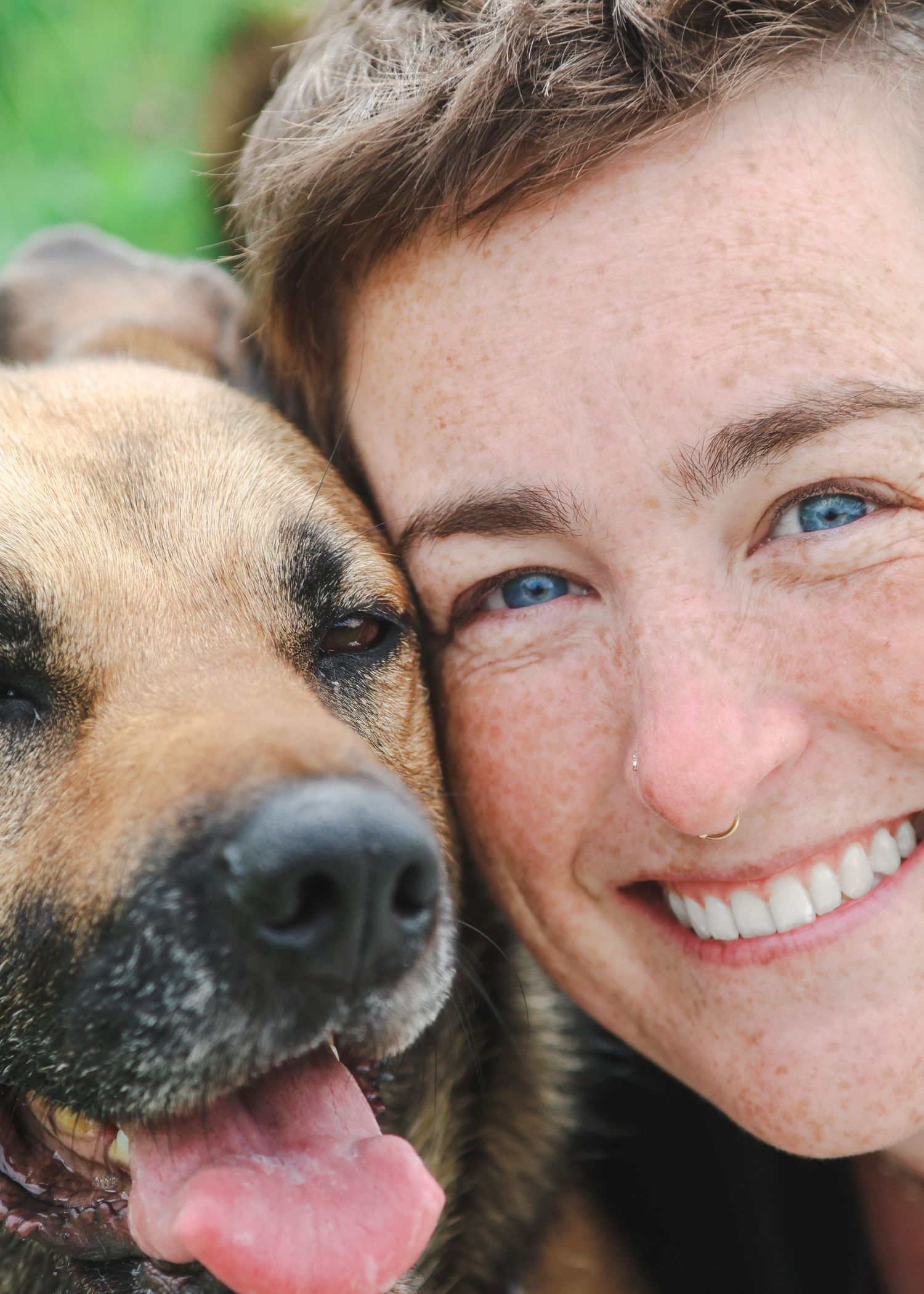 A smiling woman with blue eyes and freckles poses cheek-to-cheek with a happy German Shepherd dog outdoors.