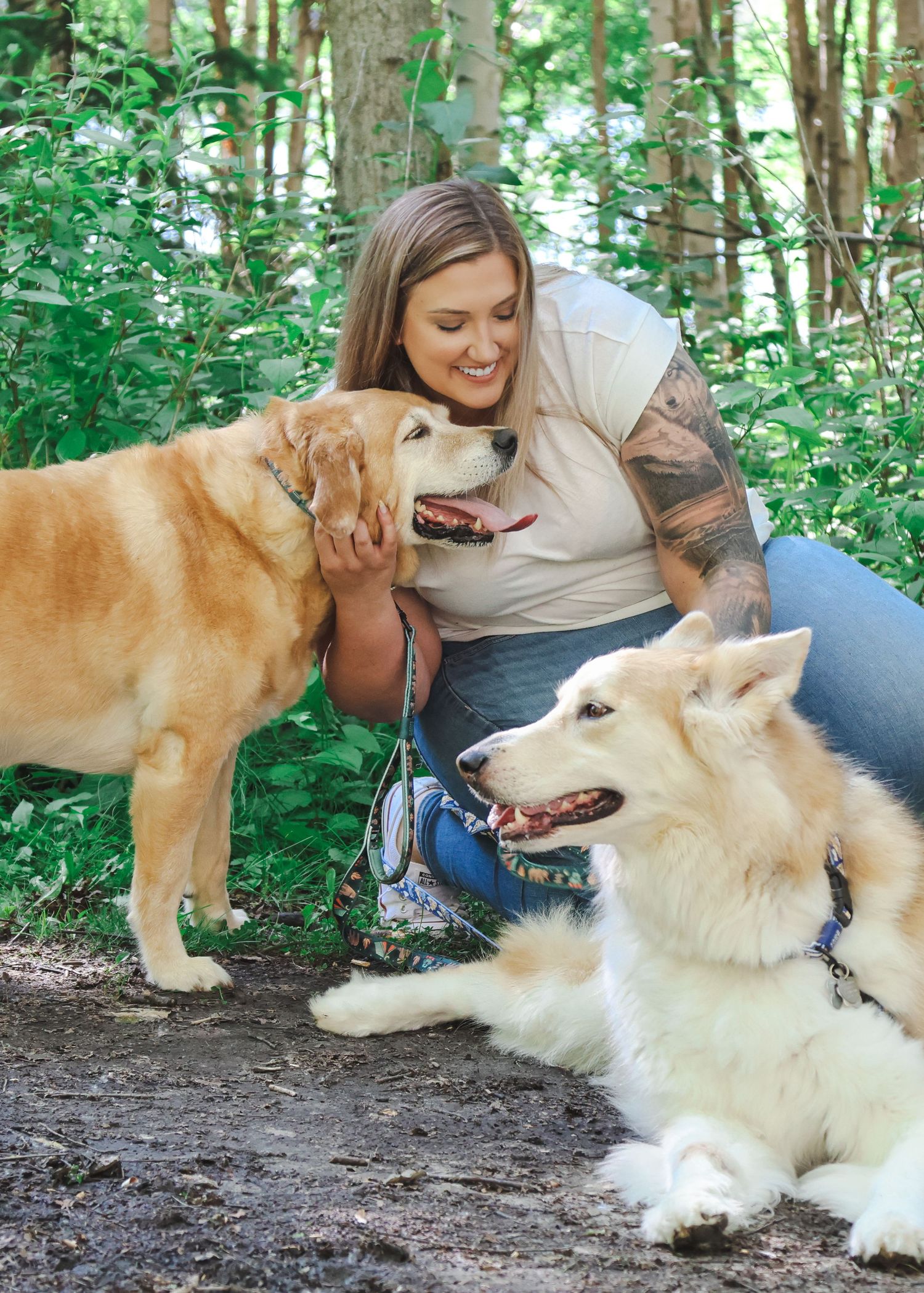 Woman with tattoos smiling and petting a golden retriever while a white fluffy dog rests nearby in a forest setting.