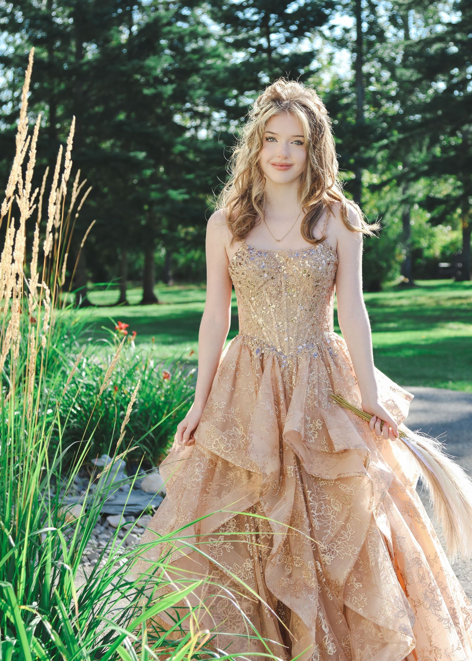 Young woman in elegant gold lace ballgown posing outdoors in lush green garden setting.