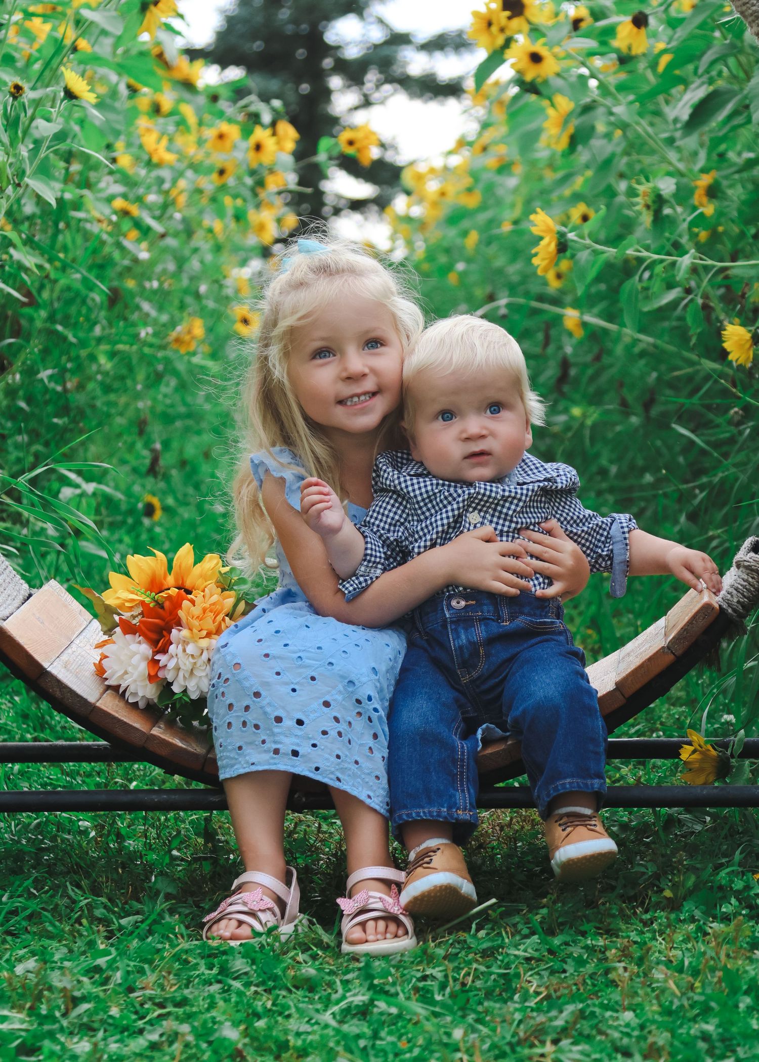 Two blonde siblings sit on a wooden bench surrounded by yellow sunflowers, the older girl hugging her baby brother.