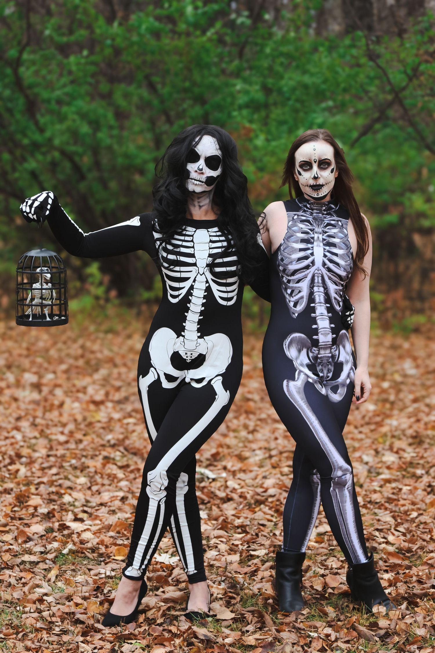 Two women in skeleton Halloween costumes with face paint pose in a forest with autumn leaves.