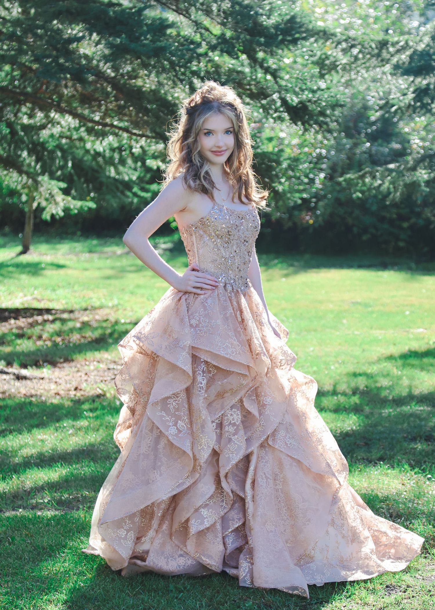 Young woman in elegant rose gold ruffled lace ball gown posing outdoors on green grass with trees.