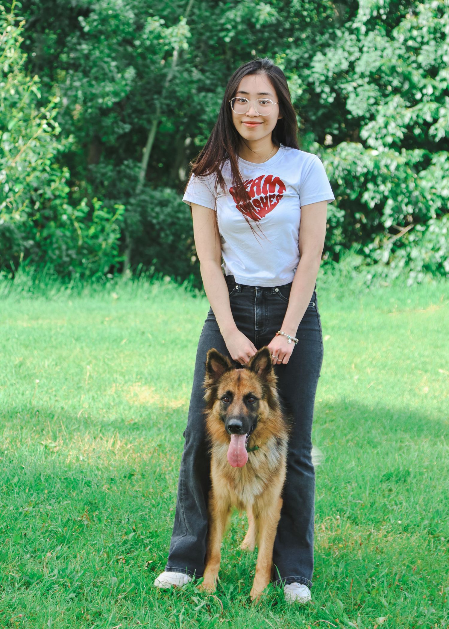 Young woman in a graphic tee and jeans stands beside a large German Shepherd on green grass.