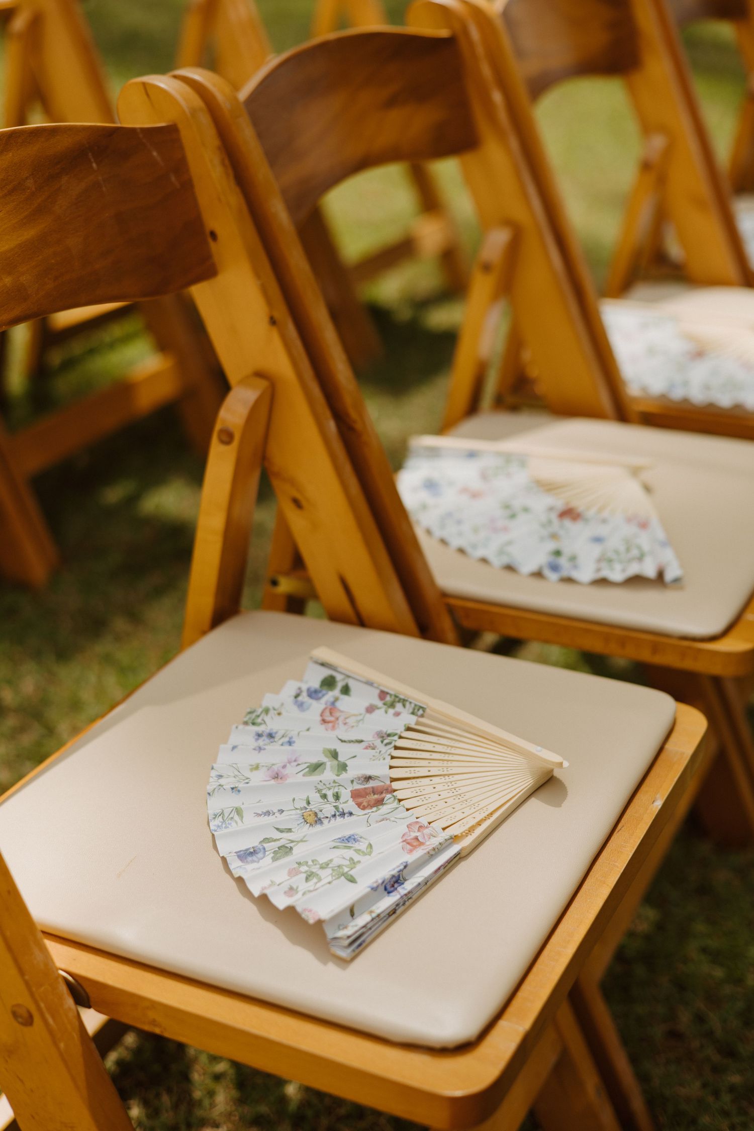 Floral paper hand fans placed on wooden folding chairs with cream cushions at an outdoor wedding ceremony.