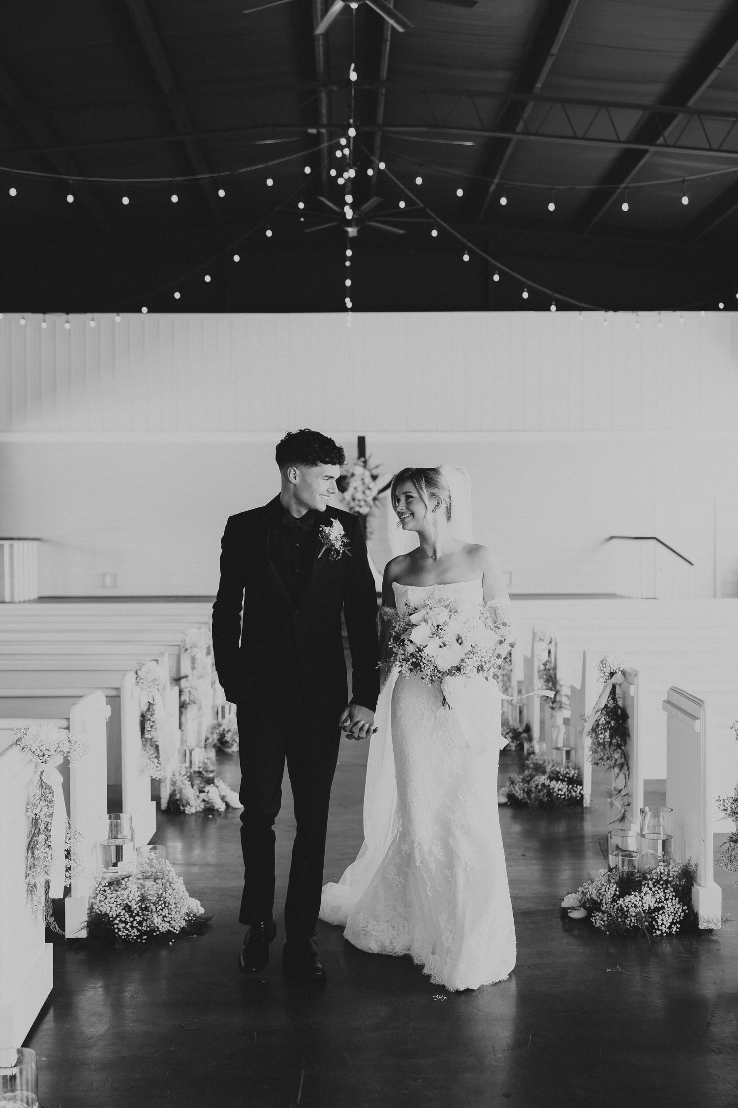 Couple walking down aisle in modern wedding venue with string lights and floral decor.