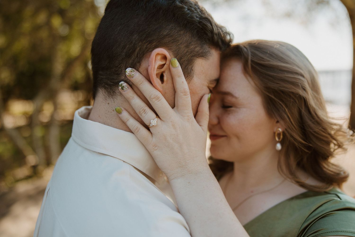 Couple sharing an intimate moment, foreheads touching, woman wearing engagement ring and green dress outdoors.
