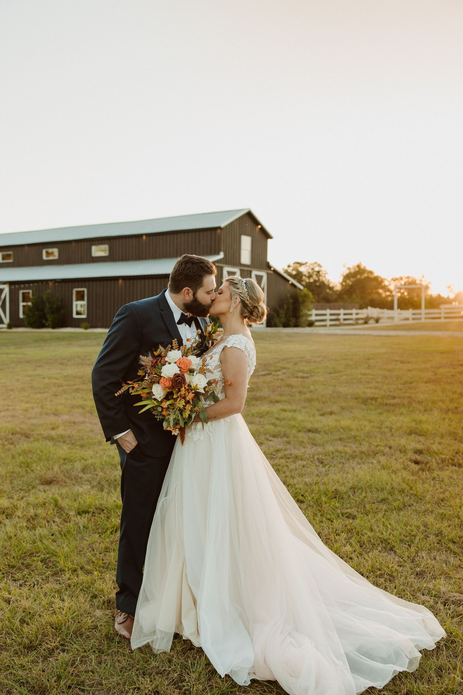 A romantic sunset wedding photo with a couple embracing in front of a rustic barn and white fence.