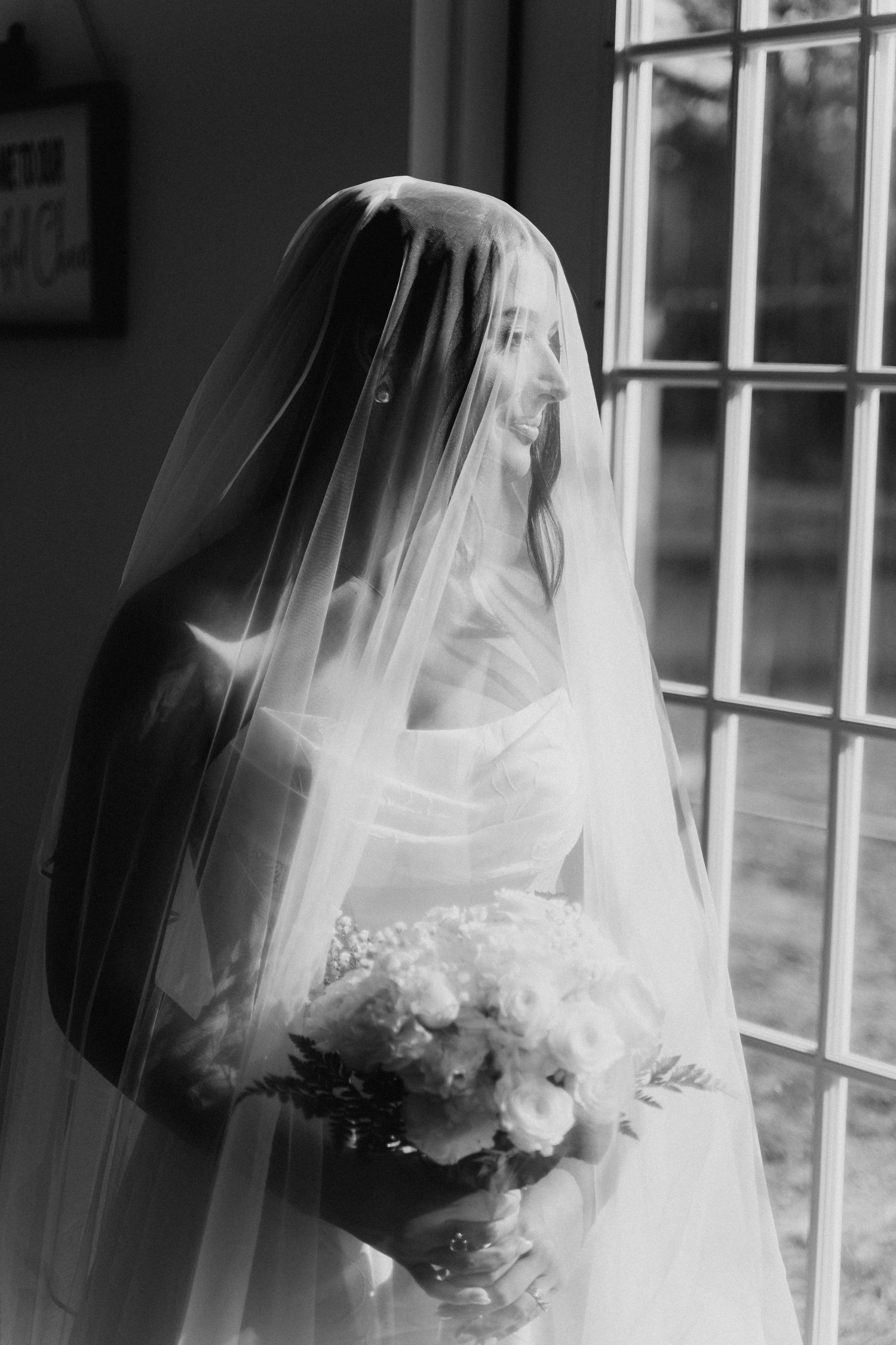 A bride in a white veil holds a bouquet of flowers while standing by a window in a black and white photograph.