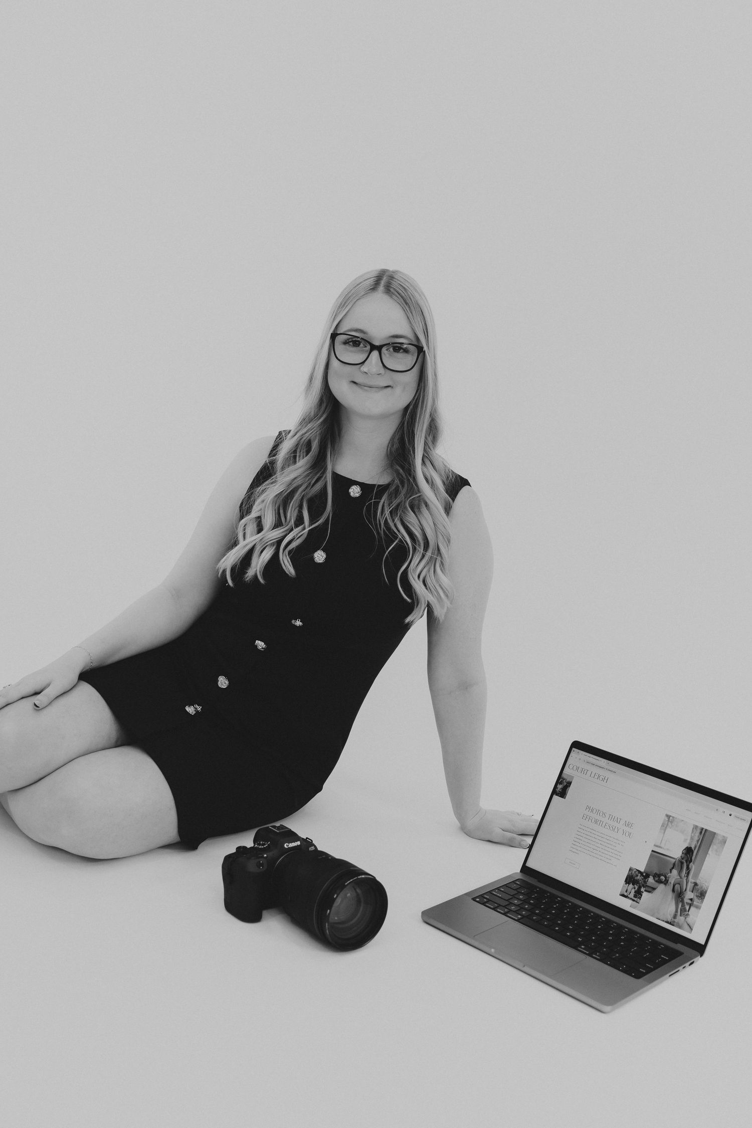 Professional photographer in a black dress with camera and laptop in minimalist studio setting.