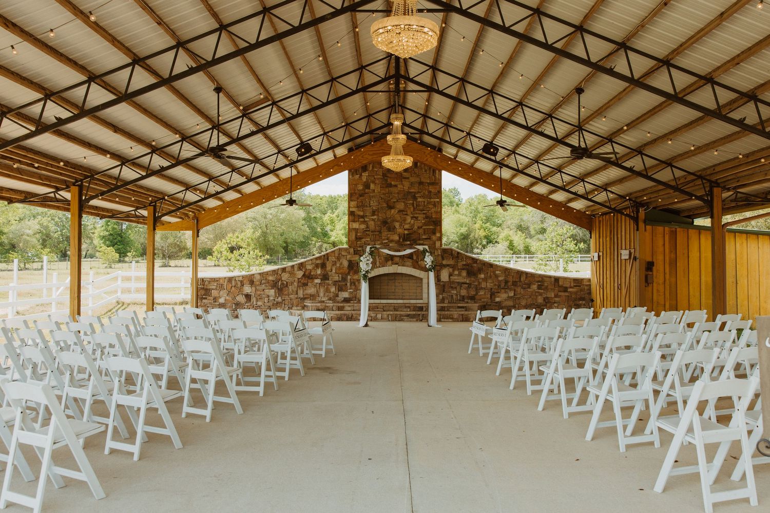 Outdoor wedding venue with white folding chairs, stone fireplace backdrop, and rustic wooden roof structure.