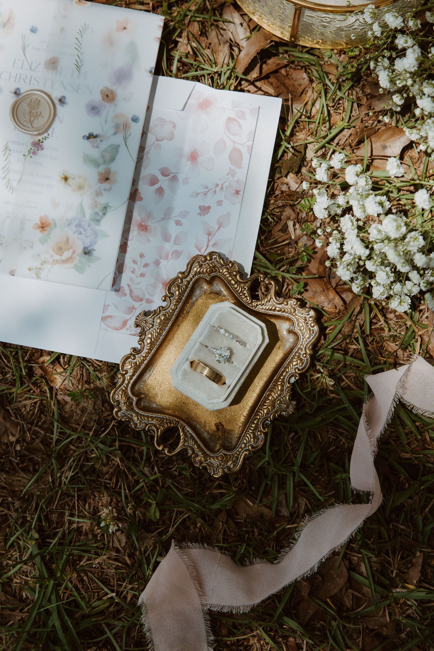 Gold ornate tray with wedding rings on grass beside floral stationery, baby's breath flowers, and silk ribbon.