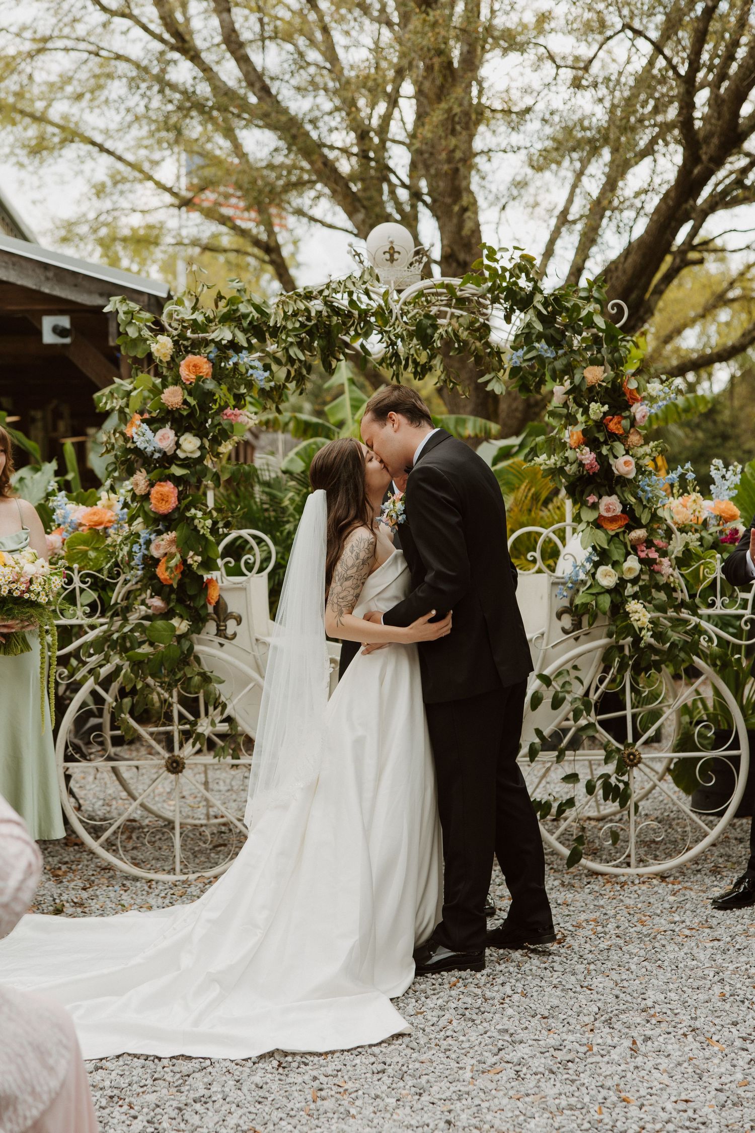 Bride and groom share first kiss at floral arch altar with colorful blooms and greenery during outdoor wedding ceremony.